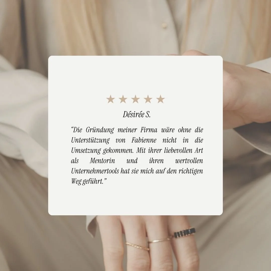 Eine Person hält ein weißes Papier mit einer 5-Sterne-Bewertung und einem Textzitat. Die Hand trägt mehrere Ringe, im Hintergrund sind beige Kleidung und Holzoberflächen sichtbar.