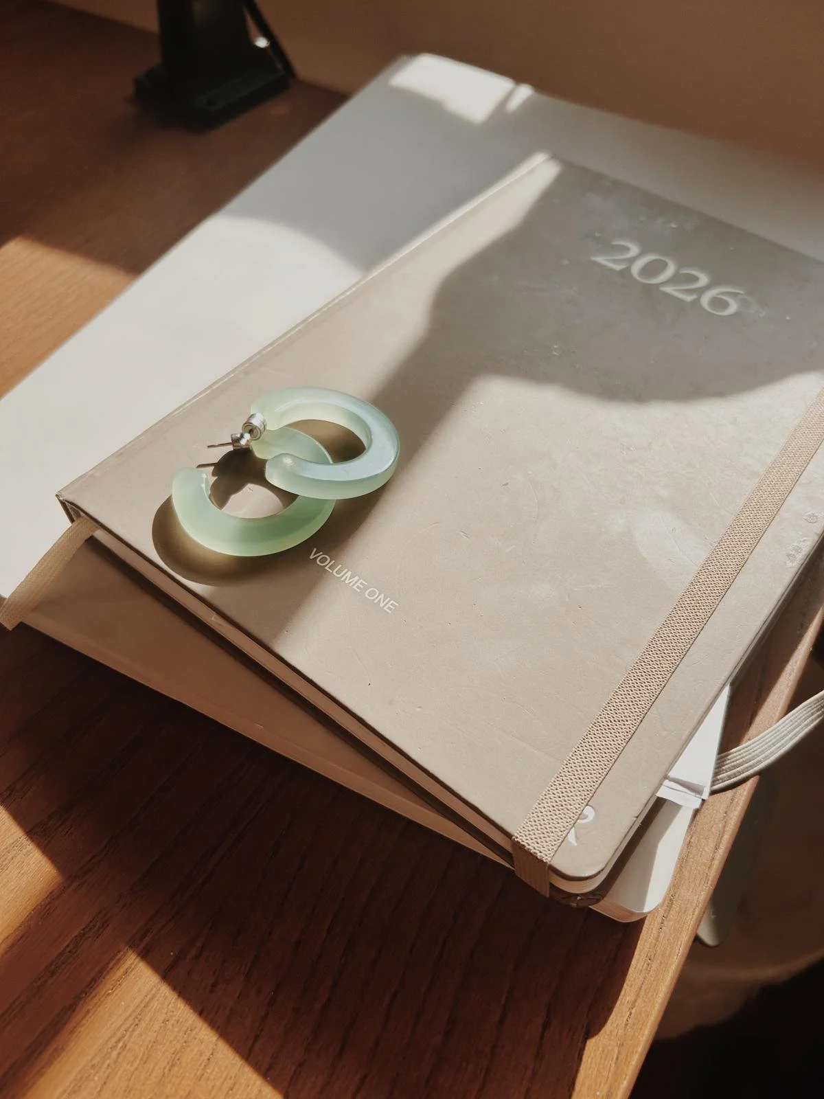 Ein offenes Notizbuch mit dem Jahr 2026 auf dem Deckblatt liegt auf einem Holztisch. Darauf liegt eine große, grüne Ohrring. Sonnenlicht fällt auf die Gegenstände und wirft Schatten.