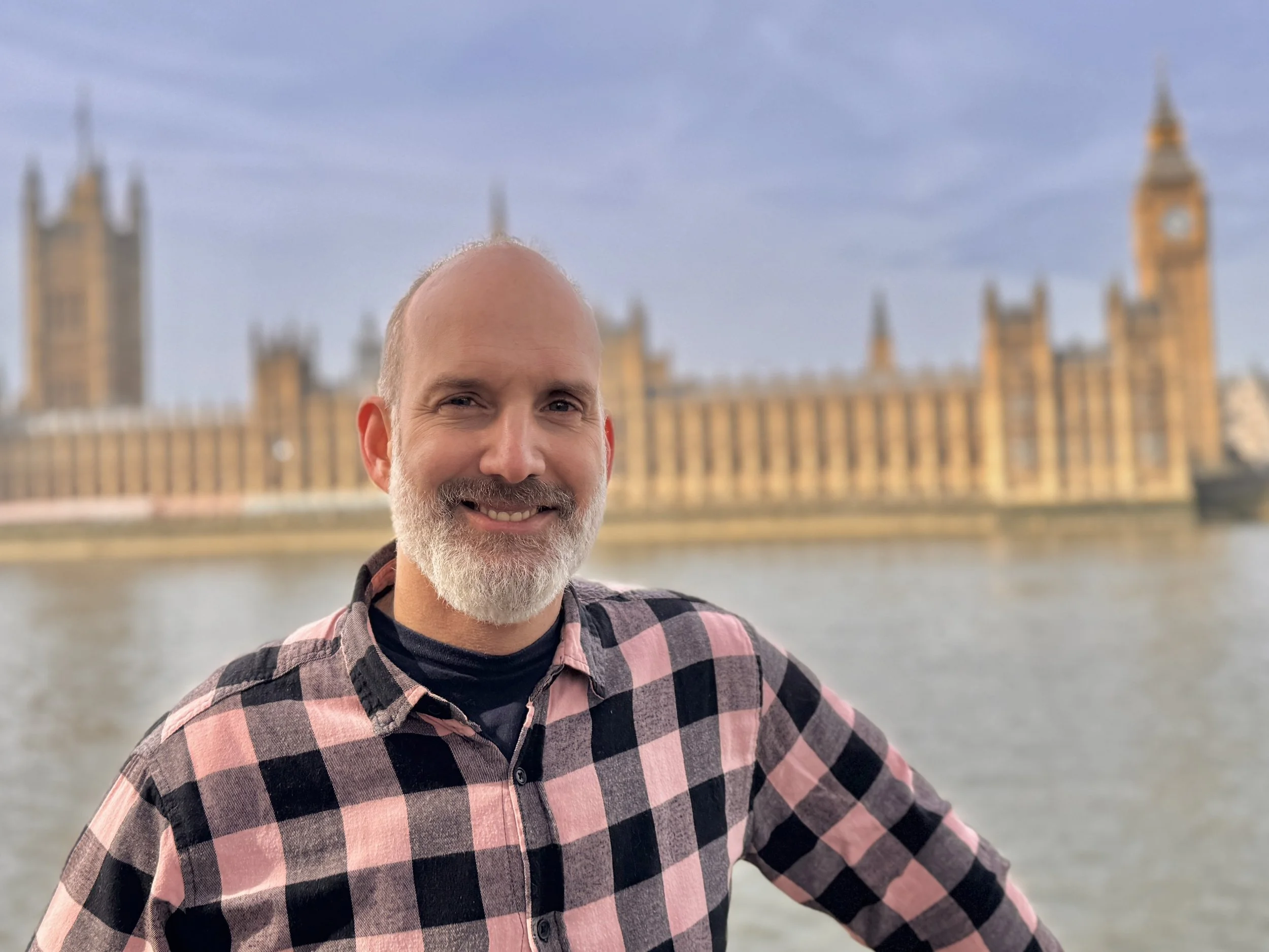 A man with a gray beard smiling in front of the UK Parliament building and Big Ben, along the river Thames.