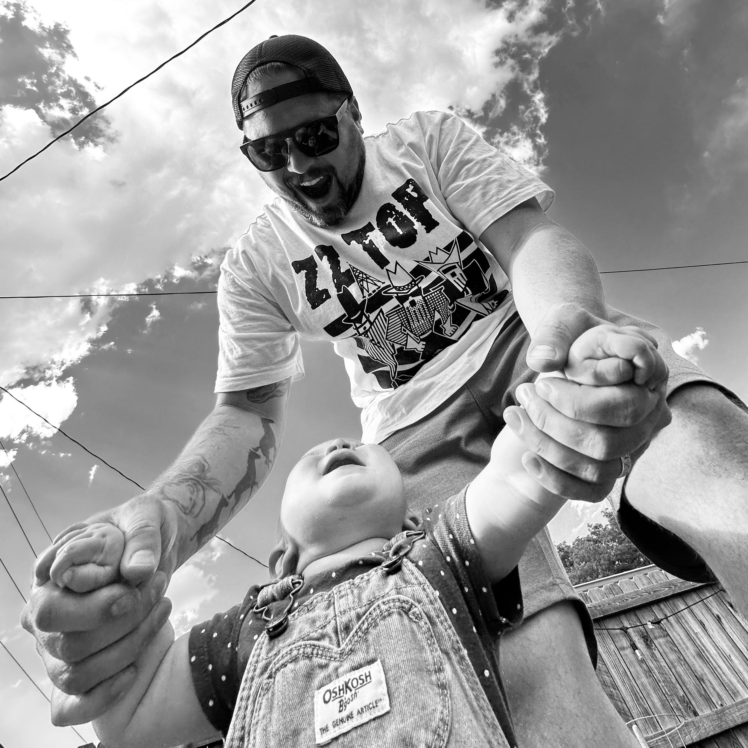 A bearded man wearing sunglasses, a baseball cap backwards, and a graphic T-shirt is playing with a young child outdoors on a sunny day with a partly cloudy sky. They are holding hands, and the man appears to be lifting or swinging the child, who is looking up.