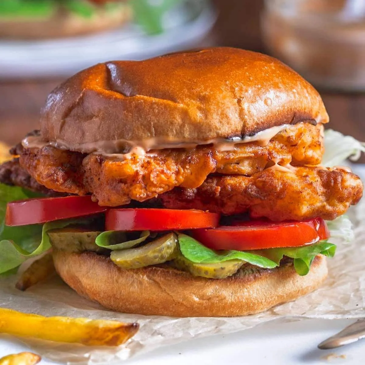 Close-up of a crispy fried chicken sandwich with lettuce, tomato slices, pickles, and mayonnaise on a toasted bun.