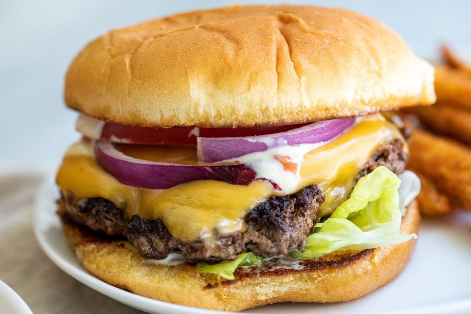 Close-up of a cheeseburger with lettuce, tomato, onions, melted cheese, and a beef patty on a bun, served with fries in the background.