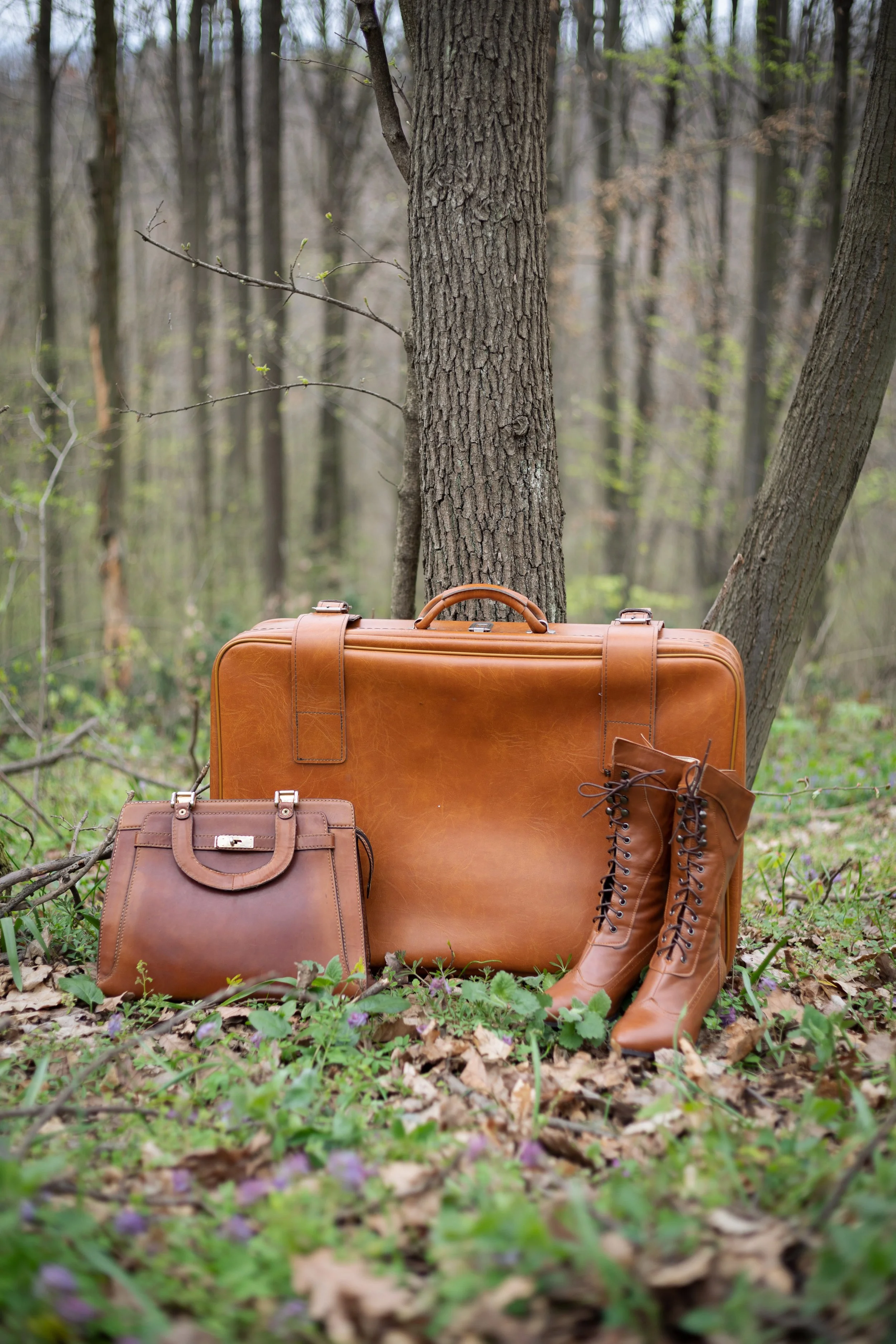 Brown leather suitcase, handbag, and lace-up boots placed on forest ground surrounded by green foliage and trees.