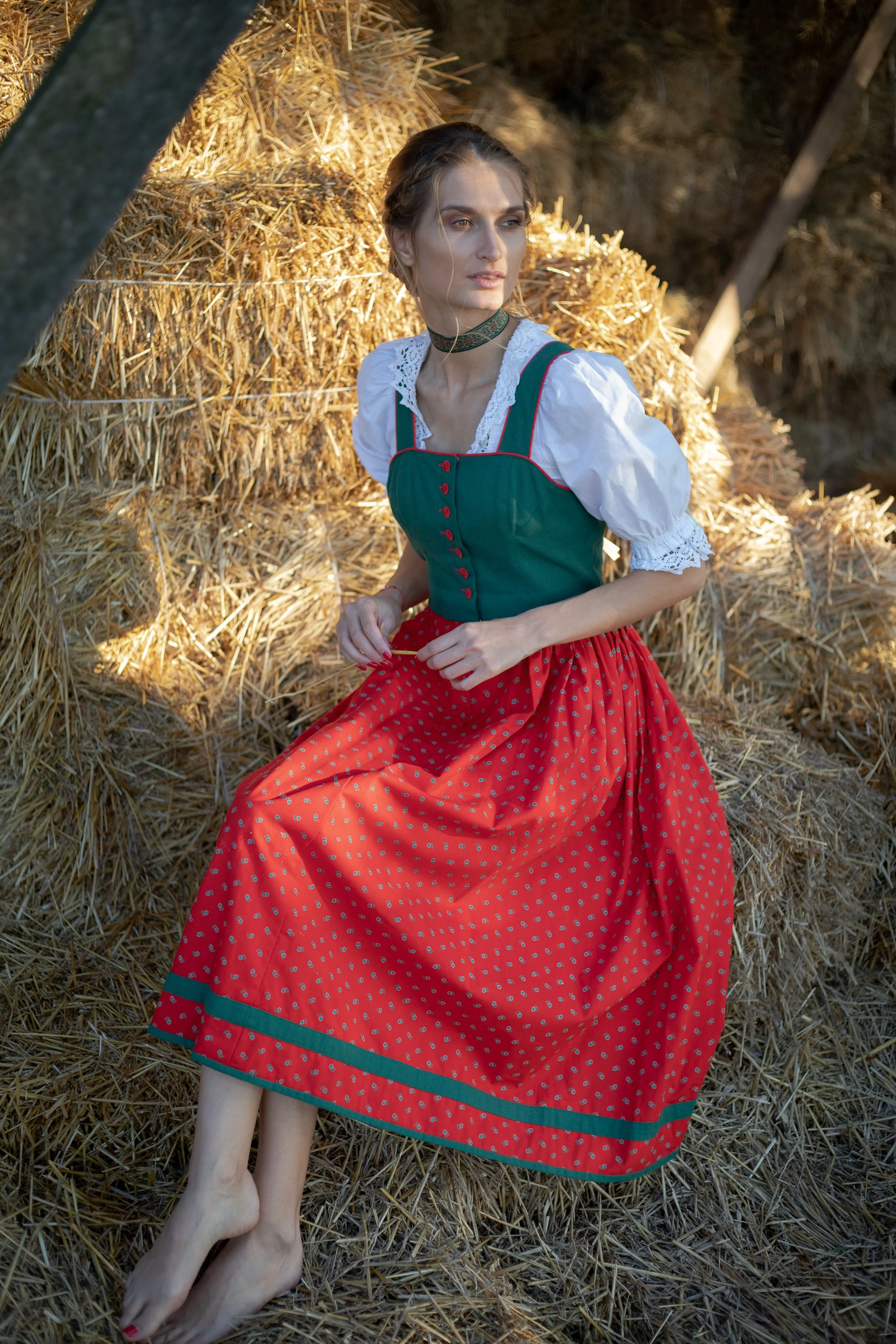 A woman dressed in a traditional European folk costume, consisting of a green bodice, white blouse, and a red skirt with a green border, sitting barefoot on hay in a barn or rural setting.
