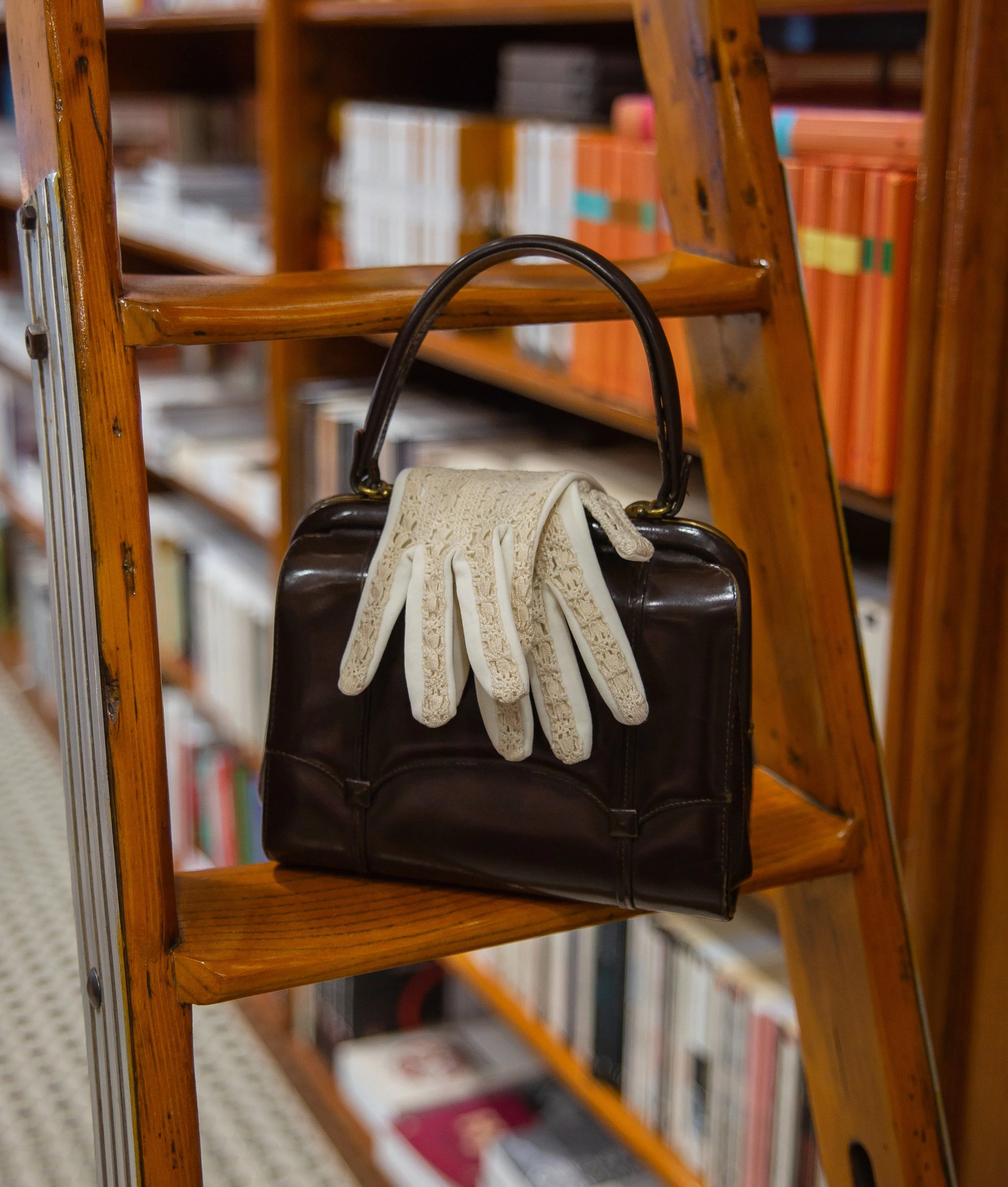 A pair of white lace gloves hanging from the handle of a black leather handbag on a wooden ladder shelf in a library.