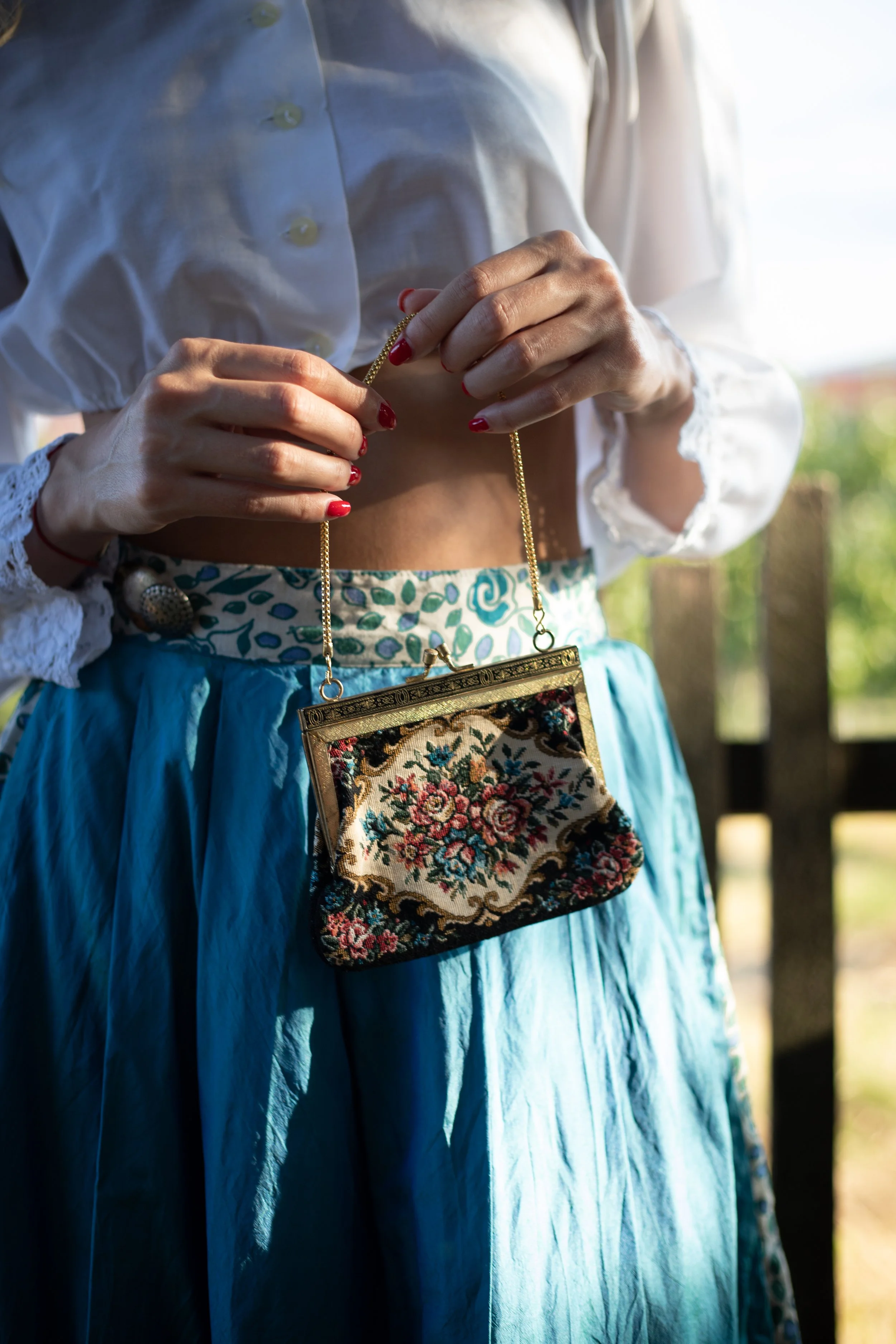A woman is holding a vintage floral fabric purse with a gold frame chain. She is wearing a white blouse with ruffled cuffs and a high-waisted blue skirt with a floral pattern.