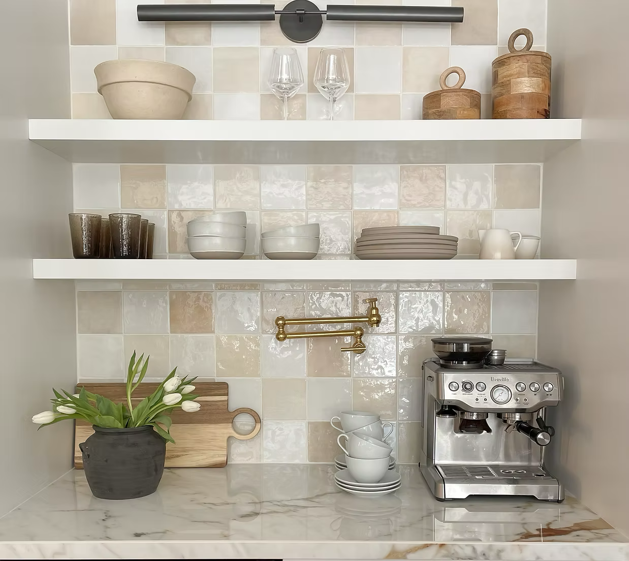 Kitchen shelf display with bowls, glasses, wooden containers, mugs, a potted plant, a cutting board, and a coffee machine. Marble countertop and tiled back wall.