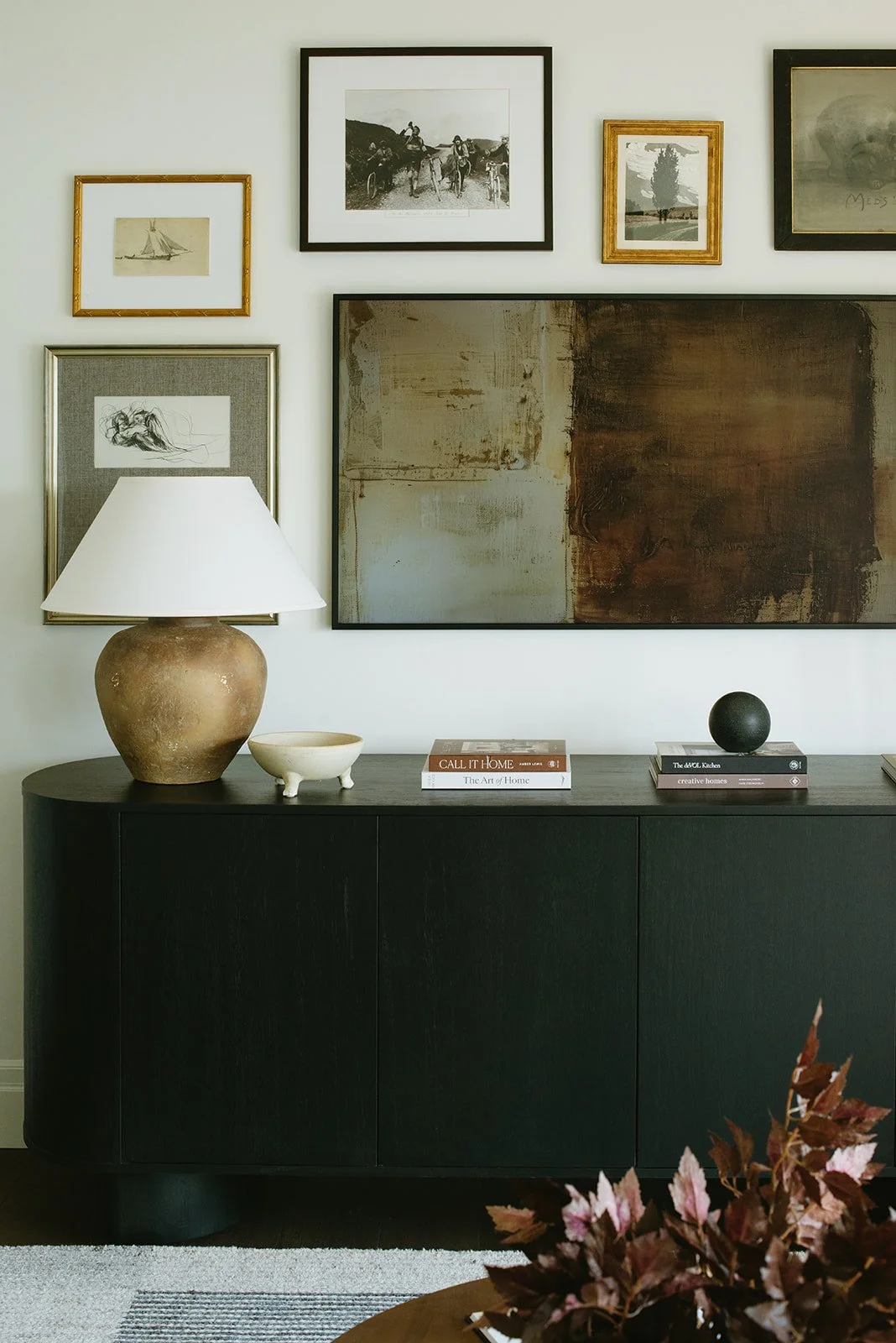 A black sideboard with a large beige ceramic table lamp, a small white ceramic bowl, and several books on top. There is a black decorative sphere on the right. The wall above features framed artwork and photographs in various sizes.
