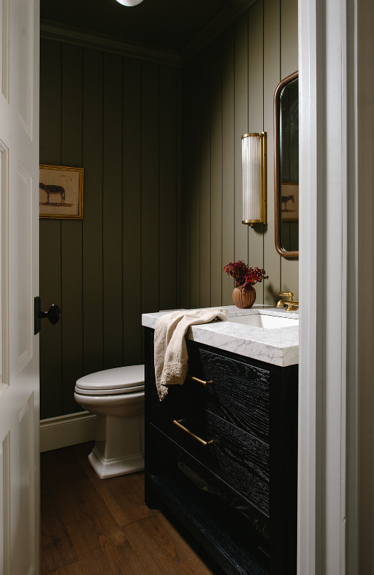 A small bathroom with dark green paneled walls, a white toilet, a black vanity with a marble countertop, a small vase of red flowers, and a mirror and wall sconce lighting.