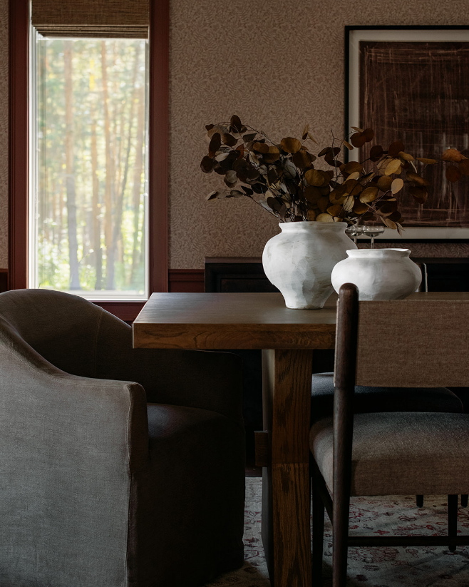 A dining room with a wooden table, surrounded by different chairs, and decorated with two white vases containing dried leaves. A window with a view of trees provides natural light.