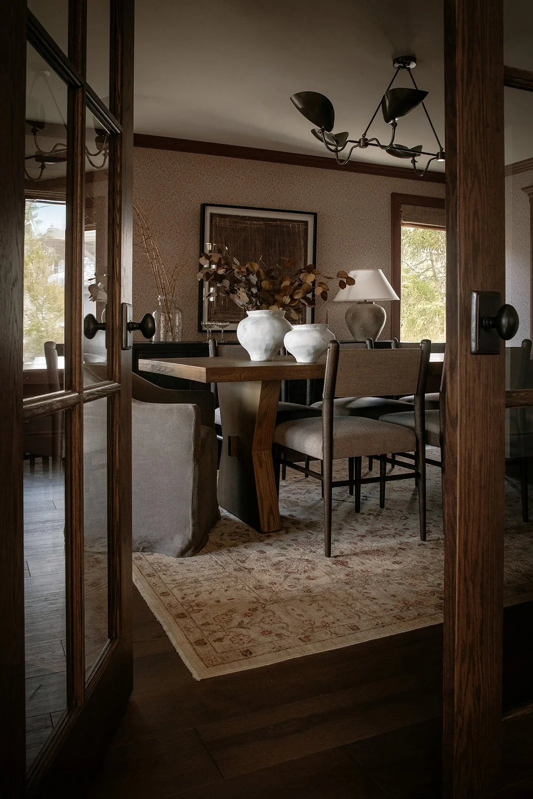 A dining room viewed through wooden French doors, featuring a rectangular wooden table, beige upholstered chairs, and a decorative rug. On the table are white vases with dried foliage, with windows and a framed artwork on the walls.
