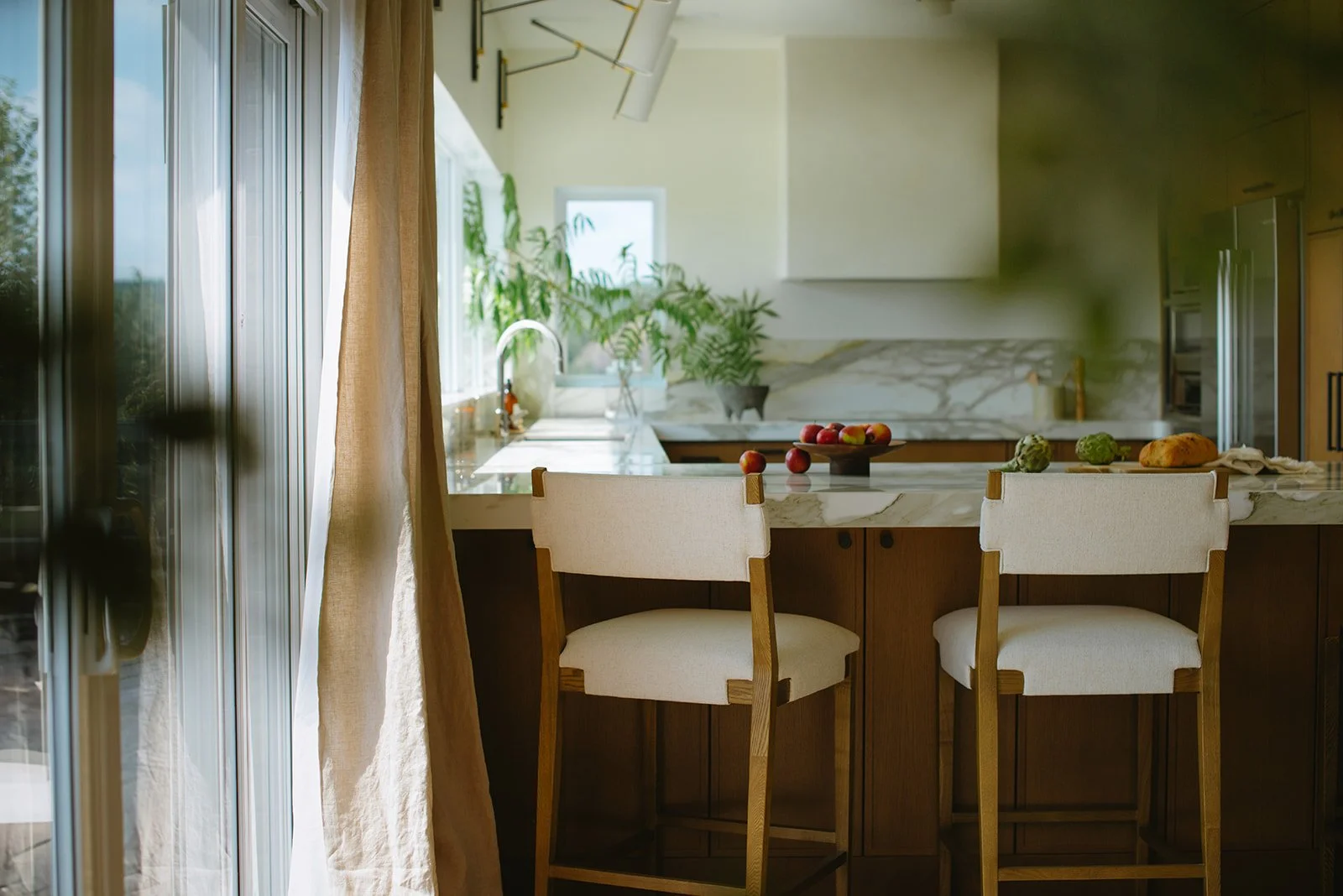 A bright kitchen with a marble island and two white chairs, apples and vegetables on the counter, a large window with curtains, and green plants in the background.