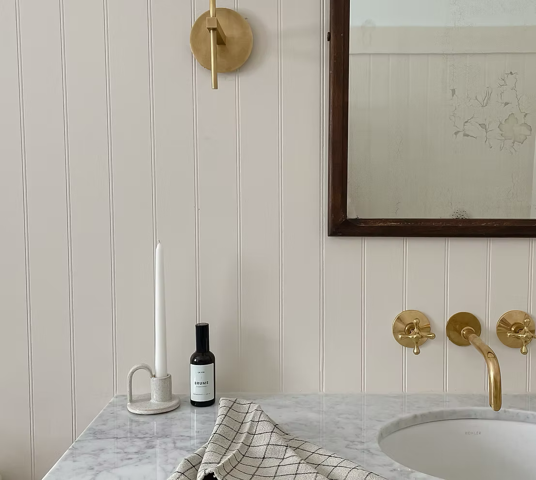 Bathroom vanity with a marble countertop, a round mirror with a wooden frame, gold wall fixtures, a candle in a holder, a bottle of soap or lotion, and a checkered towel.