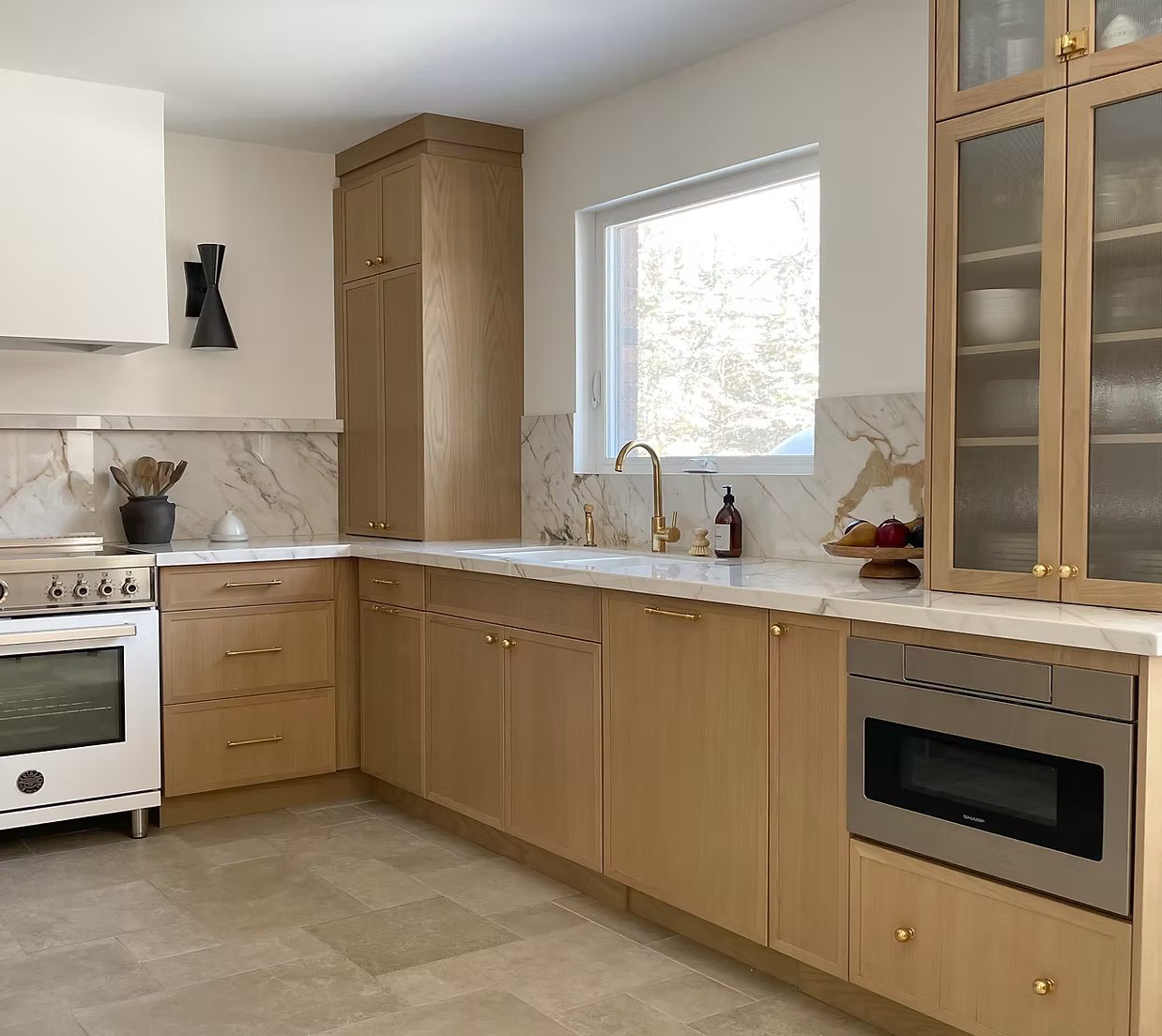 Modern kitchen with light wood cabinets, marble countertops, and stainless steel appliances, including an oven and microwave, with a window above the sink letting in natural light.