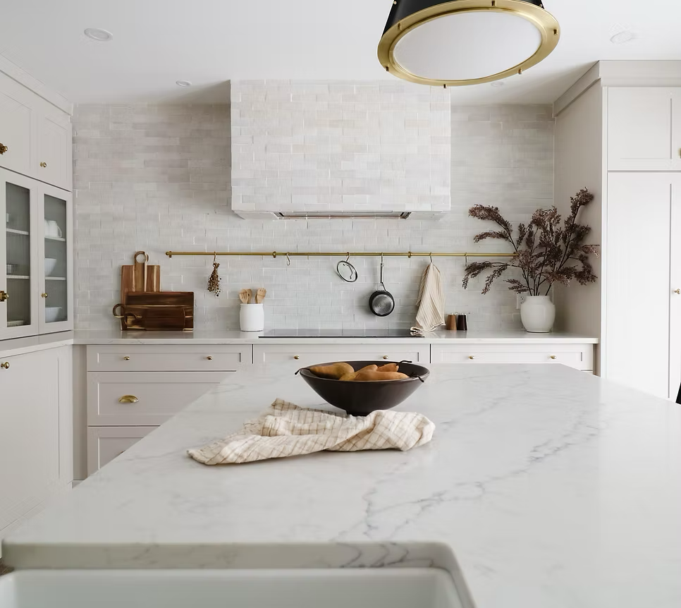 Bright white kitchen with marble countertop, white cabinets with gold hardware, hanging pots and utensils, a large white vase with dried flowers, and a black bowl with bread on the island. Large golden ceiling light fixture.
