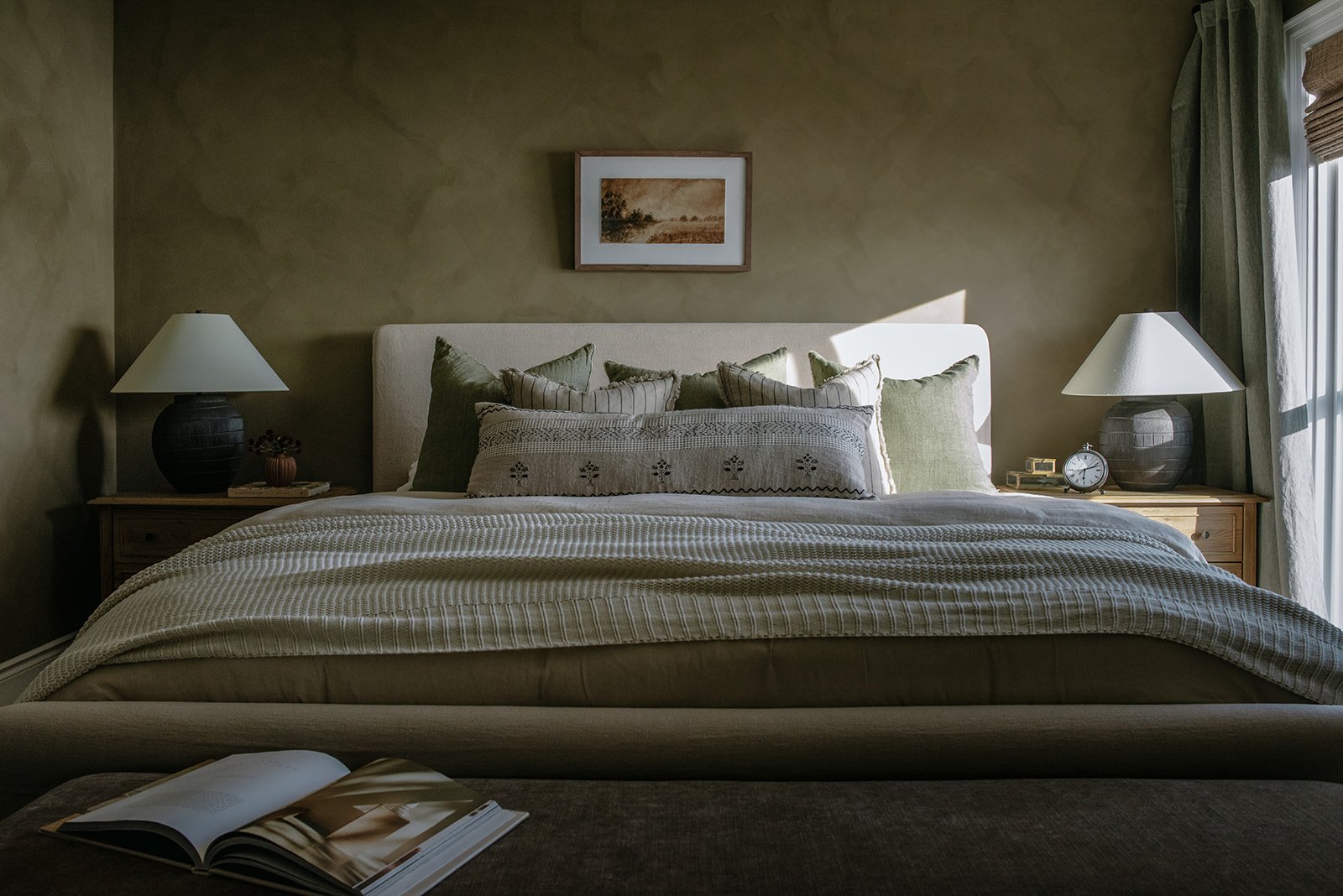 A neatly made bed with green and beige pillows in a bedroom, illuminated by sunlight from a window on the right, with side tables, lamps, a clock, and framed artwork on the wall.