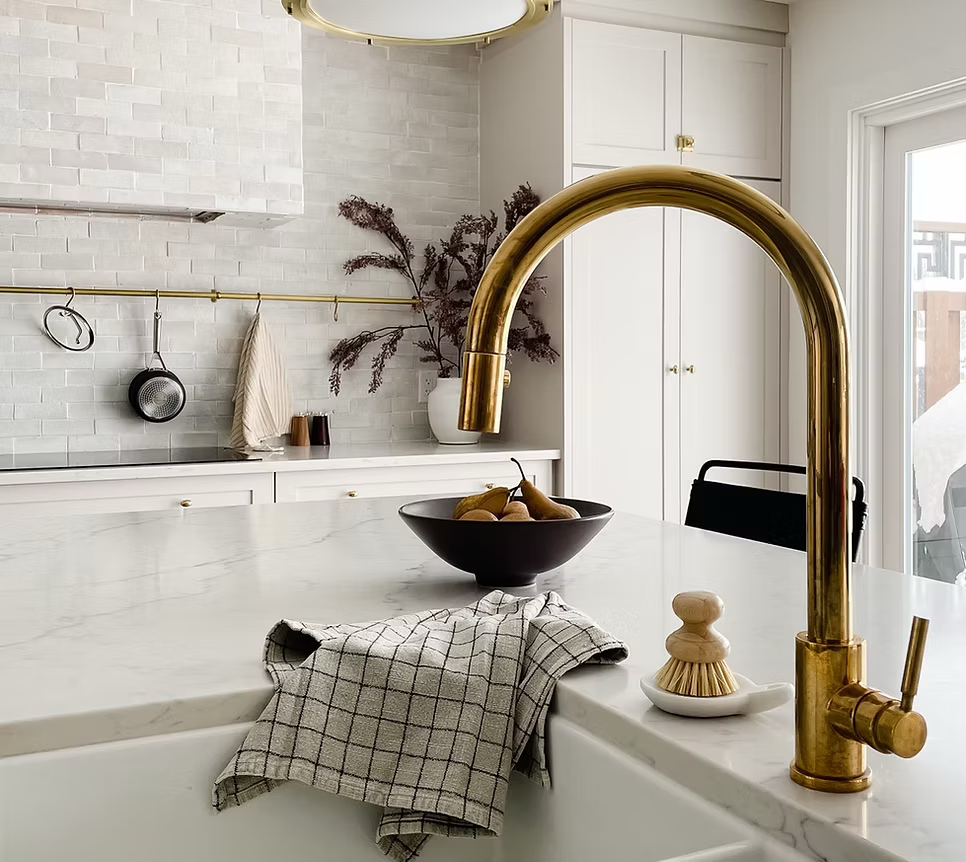 Modern kitchen with white cabinets, marble countertop, gold faucet, black bowl with pears, and a dish brush, with a window in the background.