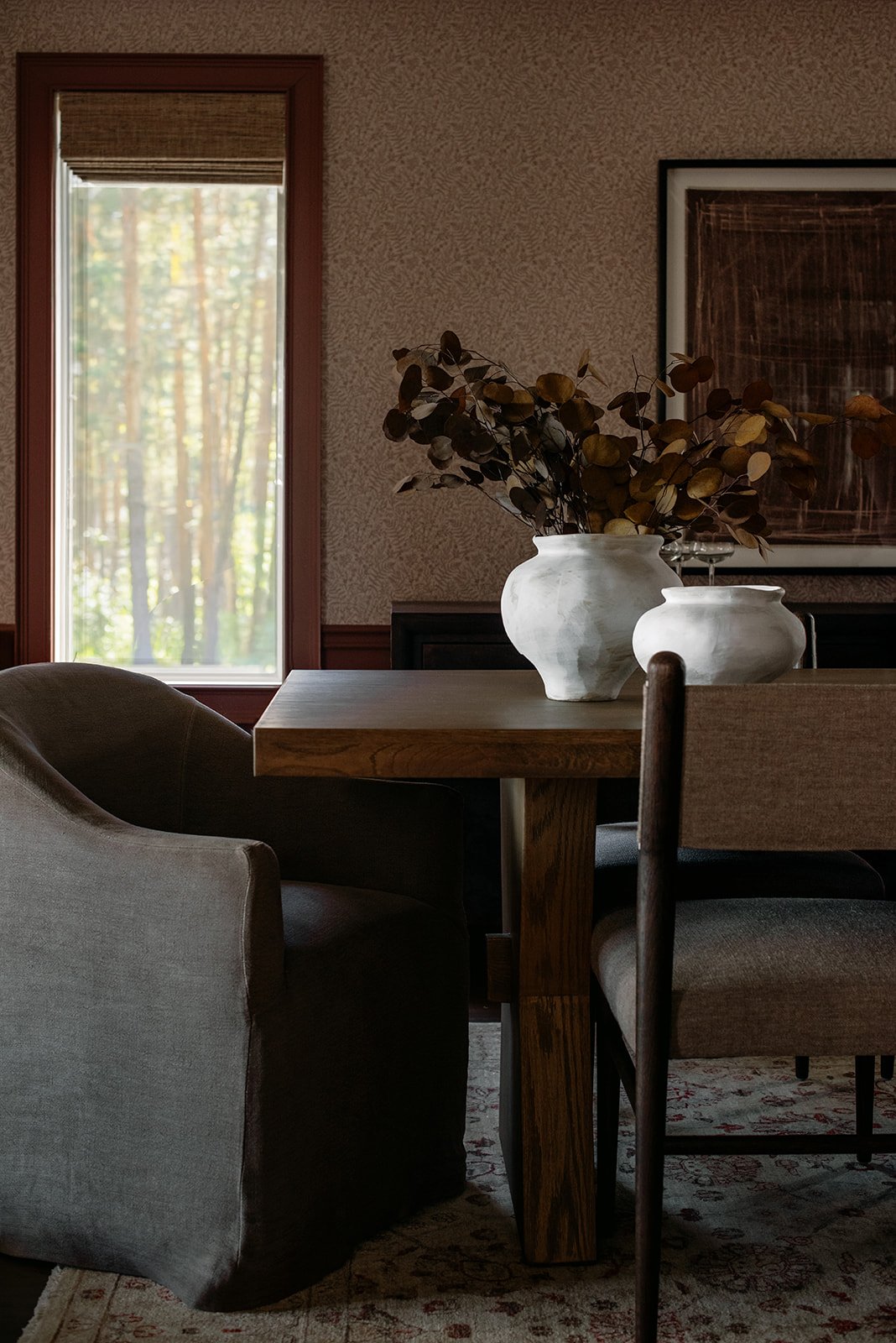 A dining room with a wooden table, gray upholstered armchair, beige fabric chairs, and decorative white vases with dried leaves on the table. A window with wooden trim and a woven blind shows trees outside. Framed artwork hangs on the wall.