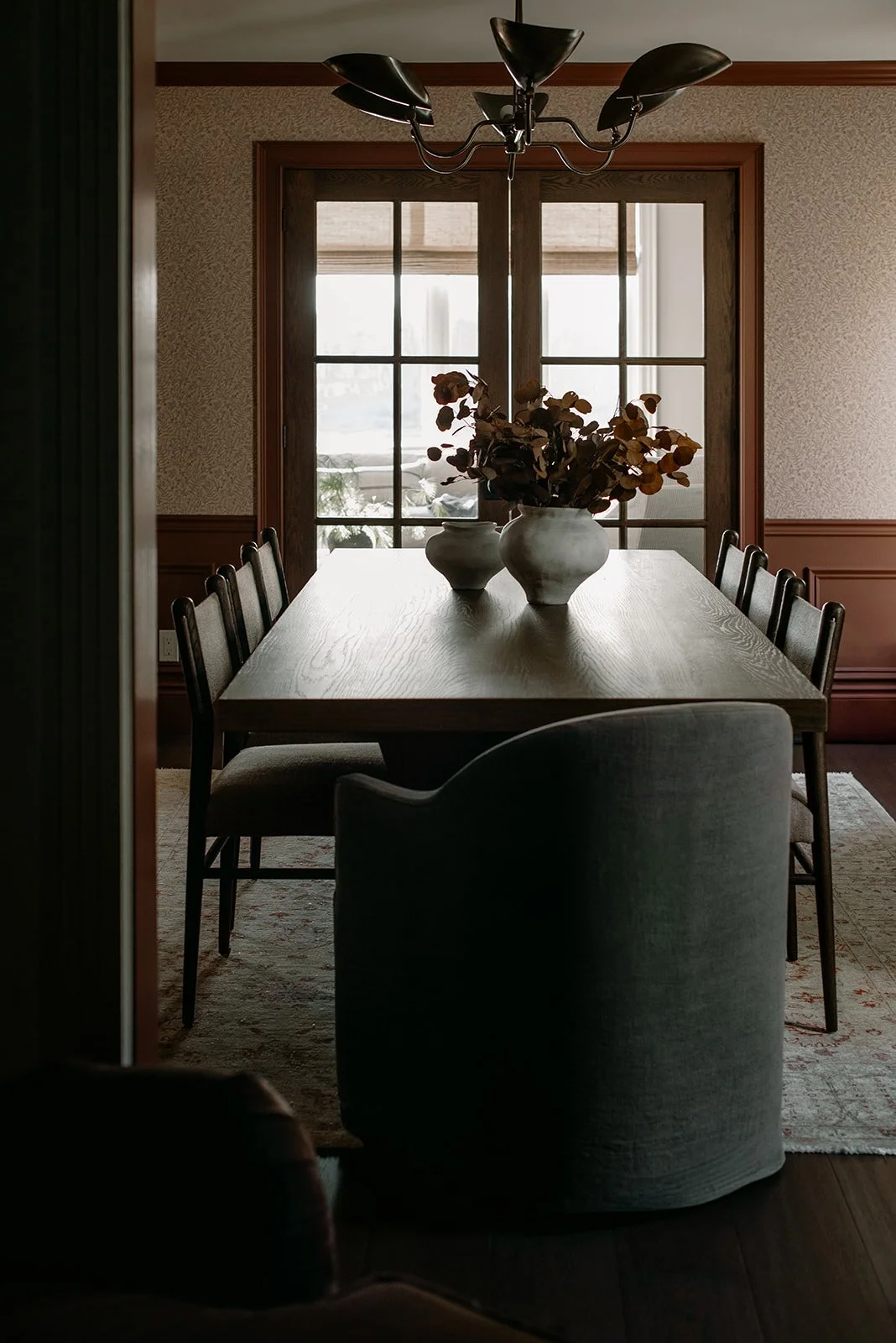 A dining room with a wooden table and eight chairs, two white vases with dried flowers on the table, a window with wooden trim, and a black chandelier overhead.