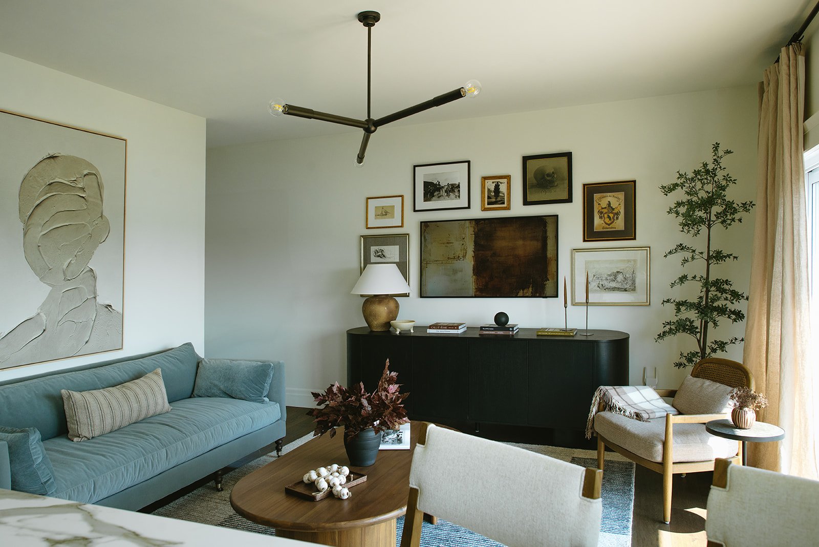 Living room with a light green sofa, a round wooden coffee table, a beige chair, a black sideboard, a large wall art piece and a gallery wall with various framed artwork, a ceiling light fixture, and a potted plant near a window with curtains.