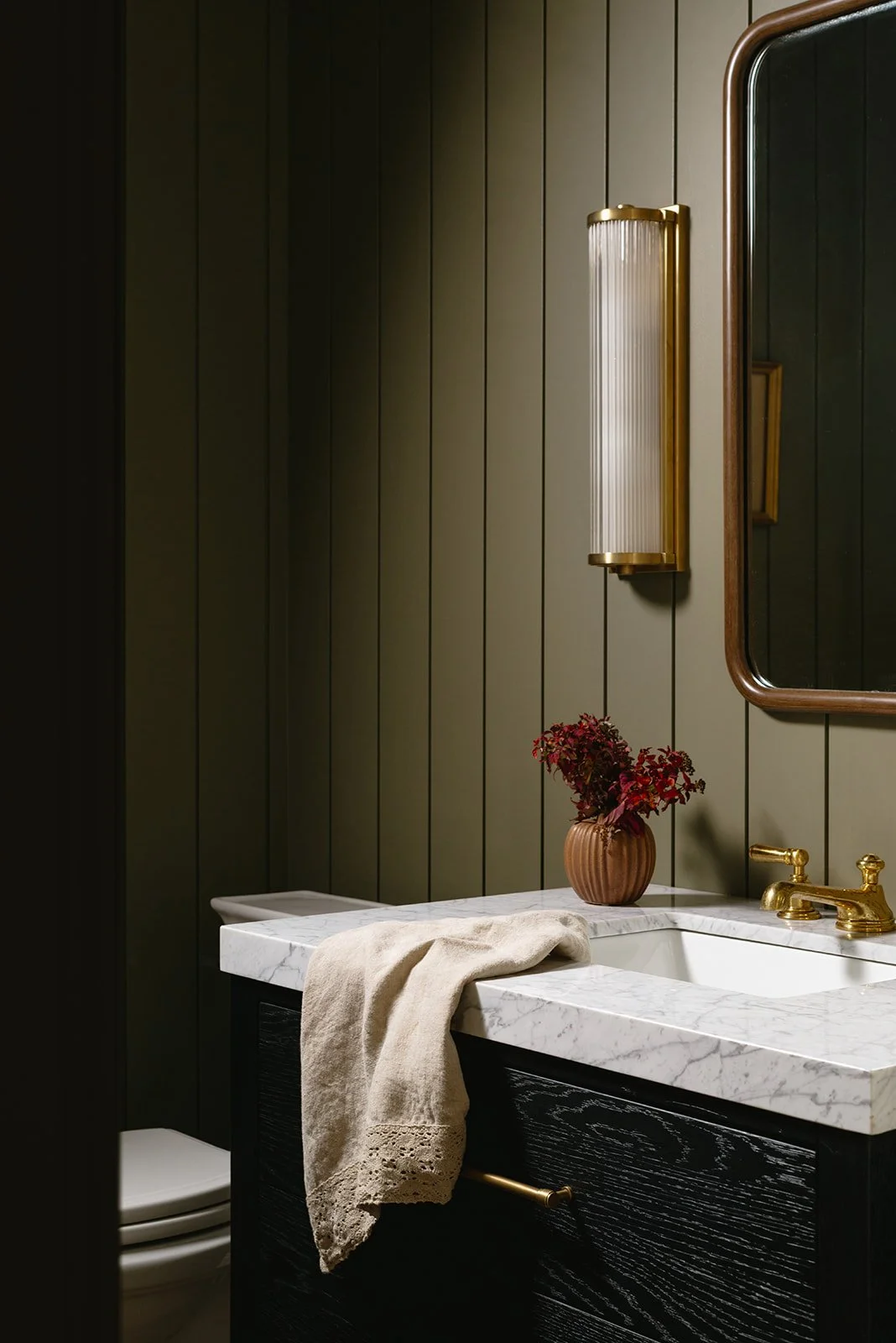 Bathroom with dark wood panel walls, marble countertop, gold fixtures, wall-mounted light, mirror, and a vase with red flowers.