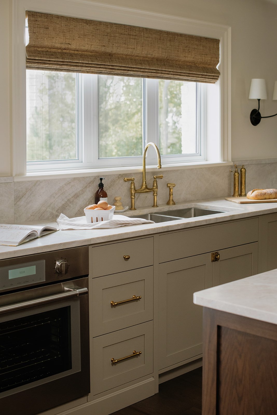 A kitchen with a window above the sink, featuring a gold faucet. The counter has a bowl of eggs, a soap dispenser, and some books. There are brass and gold decorative items on the right side of the sink. The kitchen cabinets are gray with gold handle