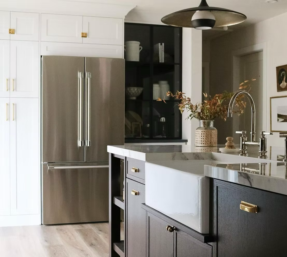 Modern kitchen with stainless steel refrigerator, white and black cabinetry, marble countertop, farmhouse sink, and decorative vase with dried flowers.