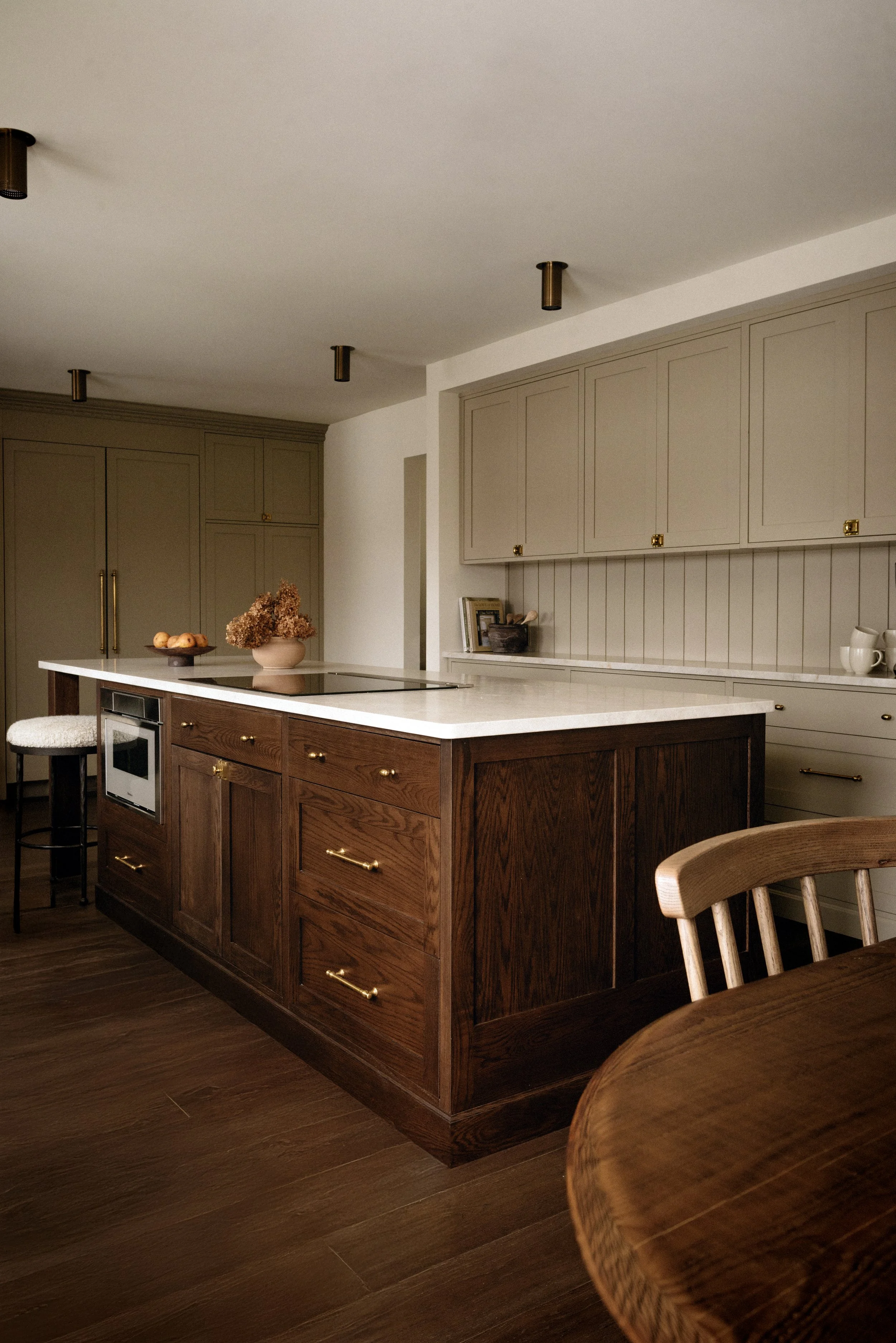 A modern kitchen with a dark wood island, beige cabinets, and a white countertop. There is a bowl of apples and a vase with dried flowers on the island, and a stool with a white cushion nearby.