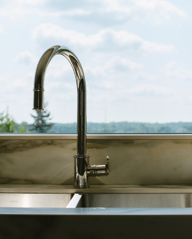 Kitchen sink with a chrome faucet against a window showing a cloudy sky and distant landscape.