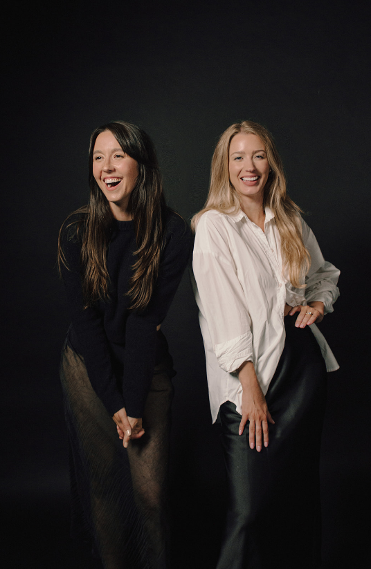 Two female interior designers in Sudbury who own LC Design Co standing side by side, smiling and looking happy, against a dark background.