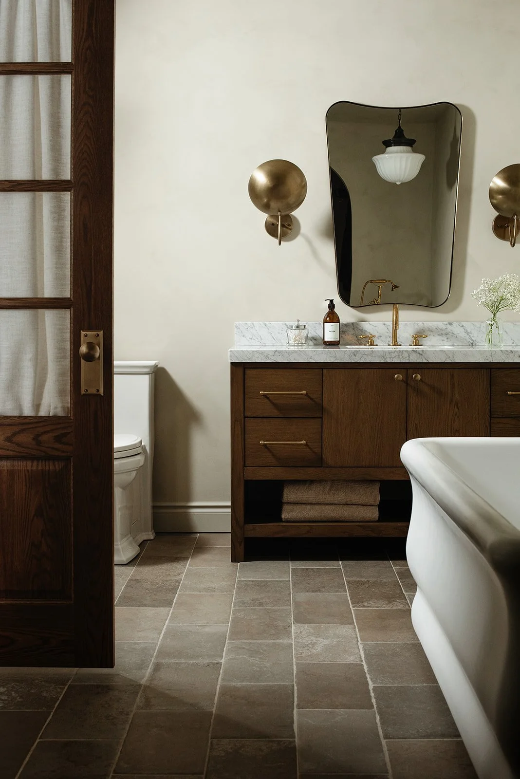 A bathroom with a wooden vanity, a marble countertop, a wall-mounted mirror, and brass wall sconces, with a white bathtub in the foreground.
