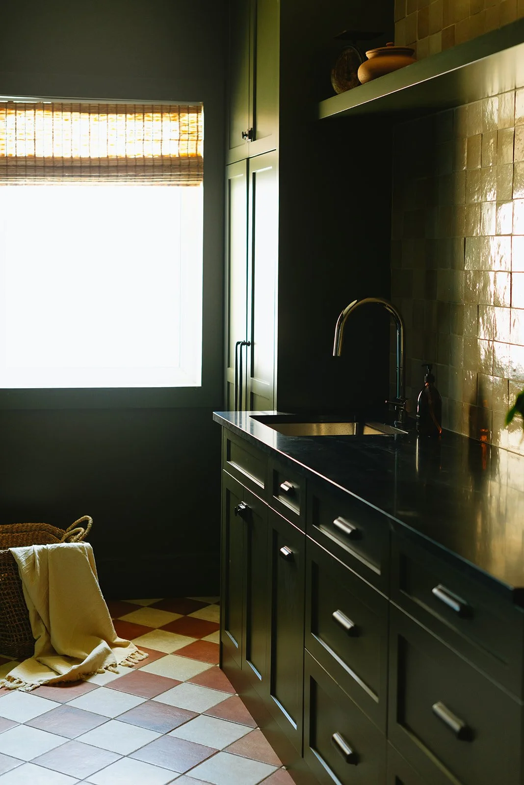 Green kitchen cabinets with black countertops, a window with bamboo shade, and a tiled floor with a yellow towel on the floor.