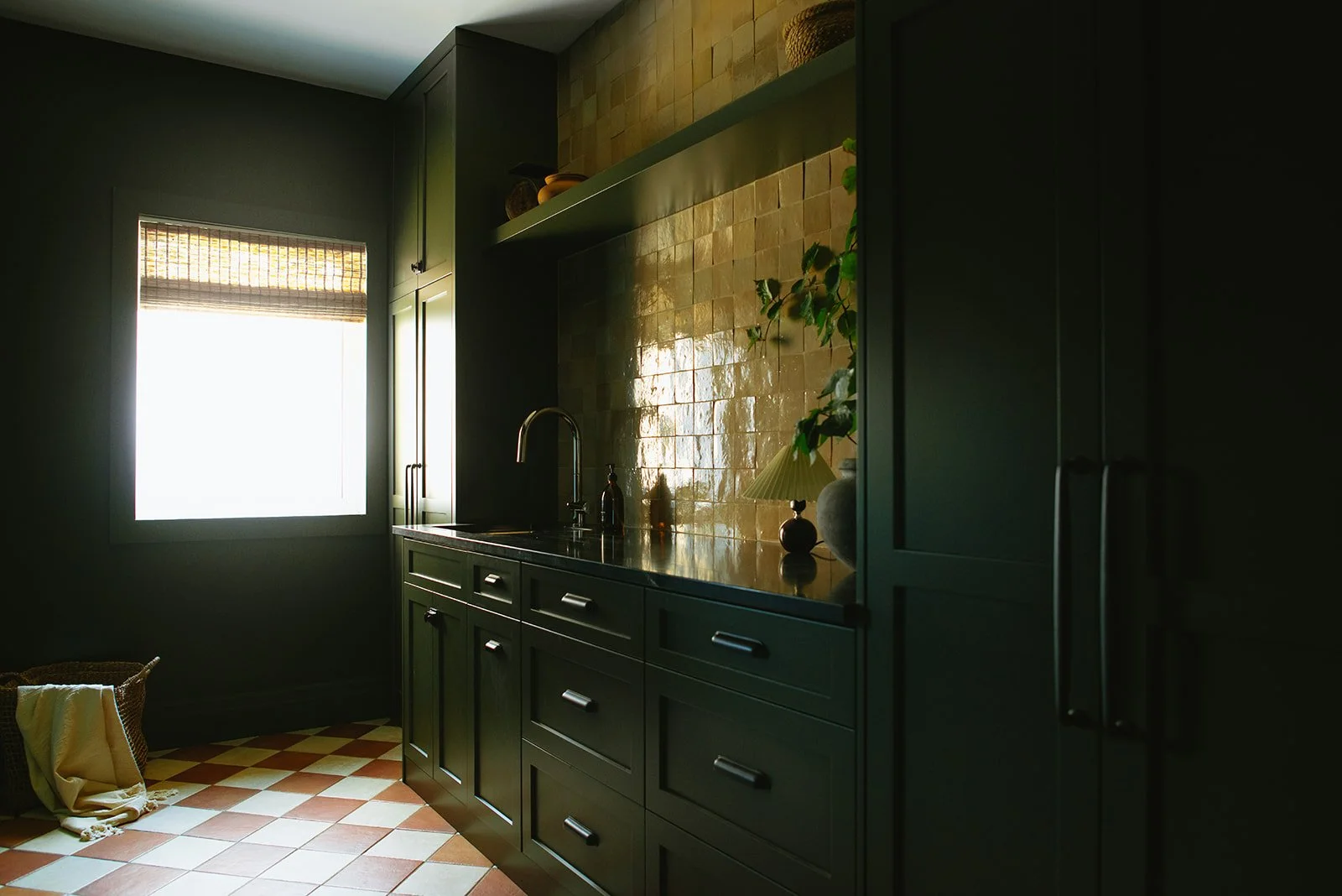 A kitchen with dark green cabinets, a window with a bamboo blind, and a mustard-colored tiled backsplash. There are decorative items on the open shelf and a plant on the counter. The floor has a checkered pattern of orange and beige tiles.