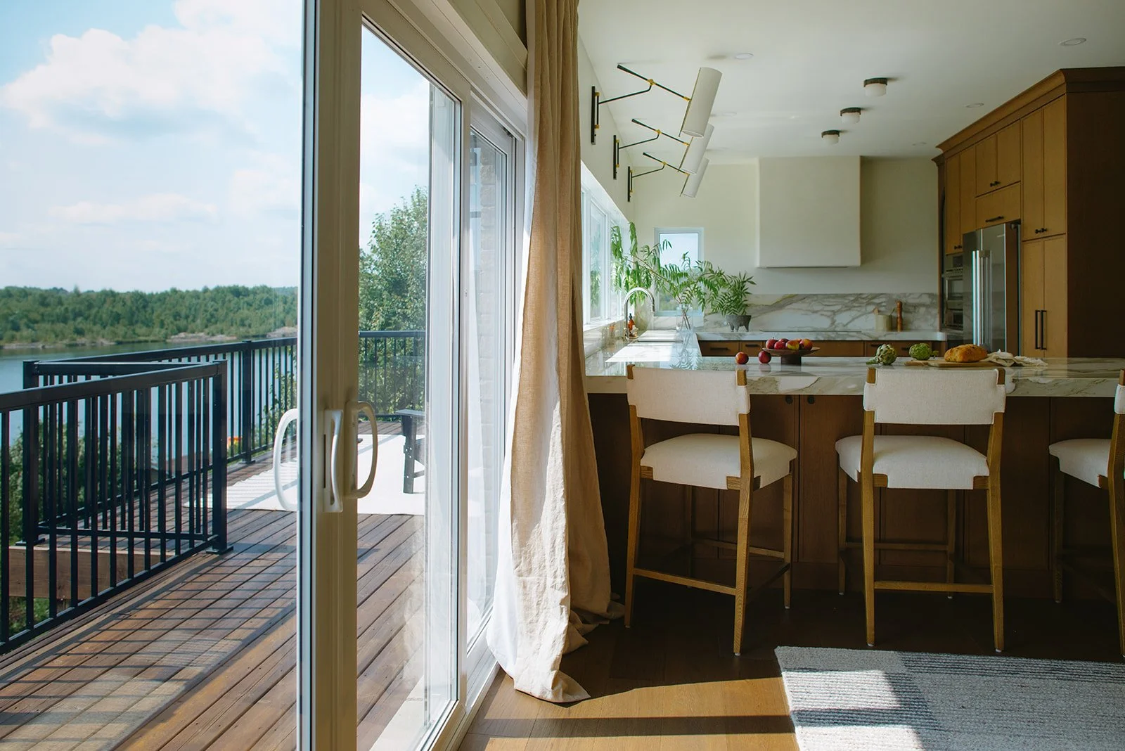 Kitchen with view of outdoor balcony with black railing, trees, and water in the distance, modern design with wooden cabinets, marble countertops, bar stools, and plants