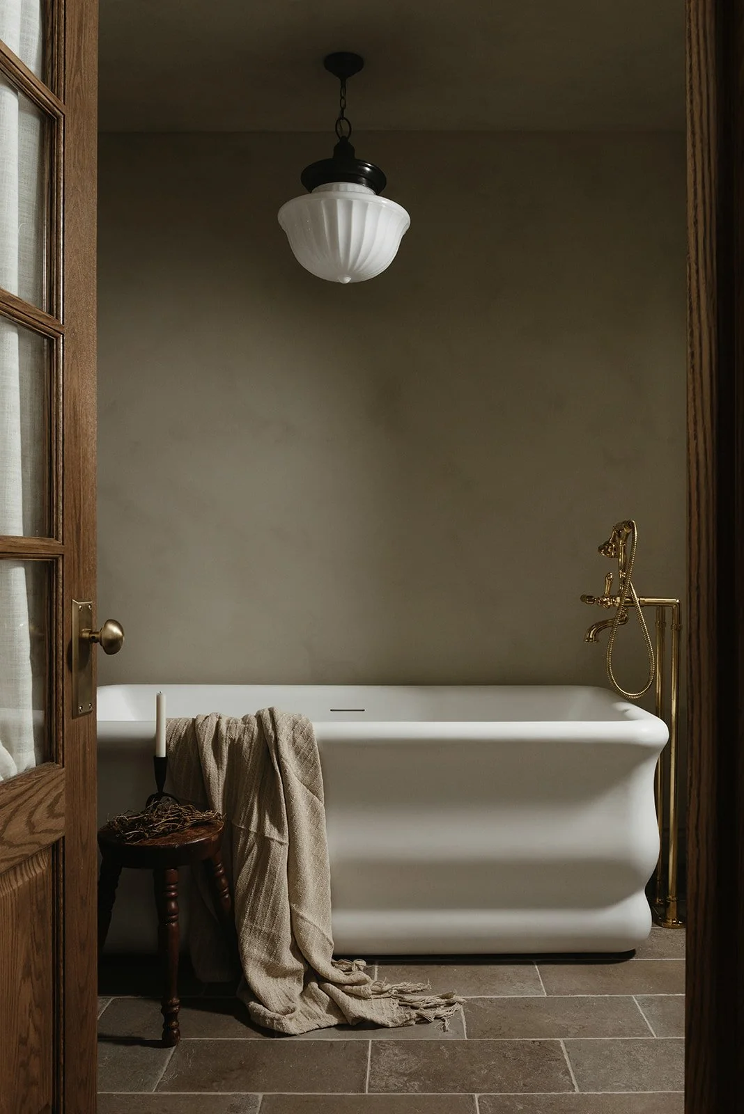 A vintage-style bathroom with a white clawfoot bathtub, a brass faucet and showerhead, a small wooden stool with a candle and a rag, and a beige towel draped over the tub. A ceiling light fixture is hanging from the ceiling, and the bathroom door is 