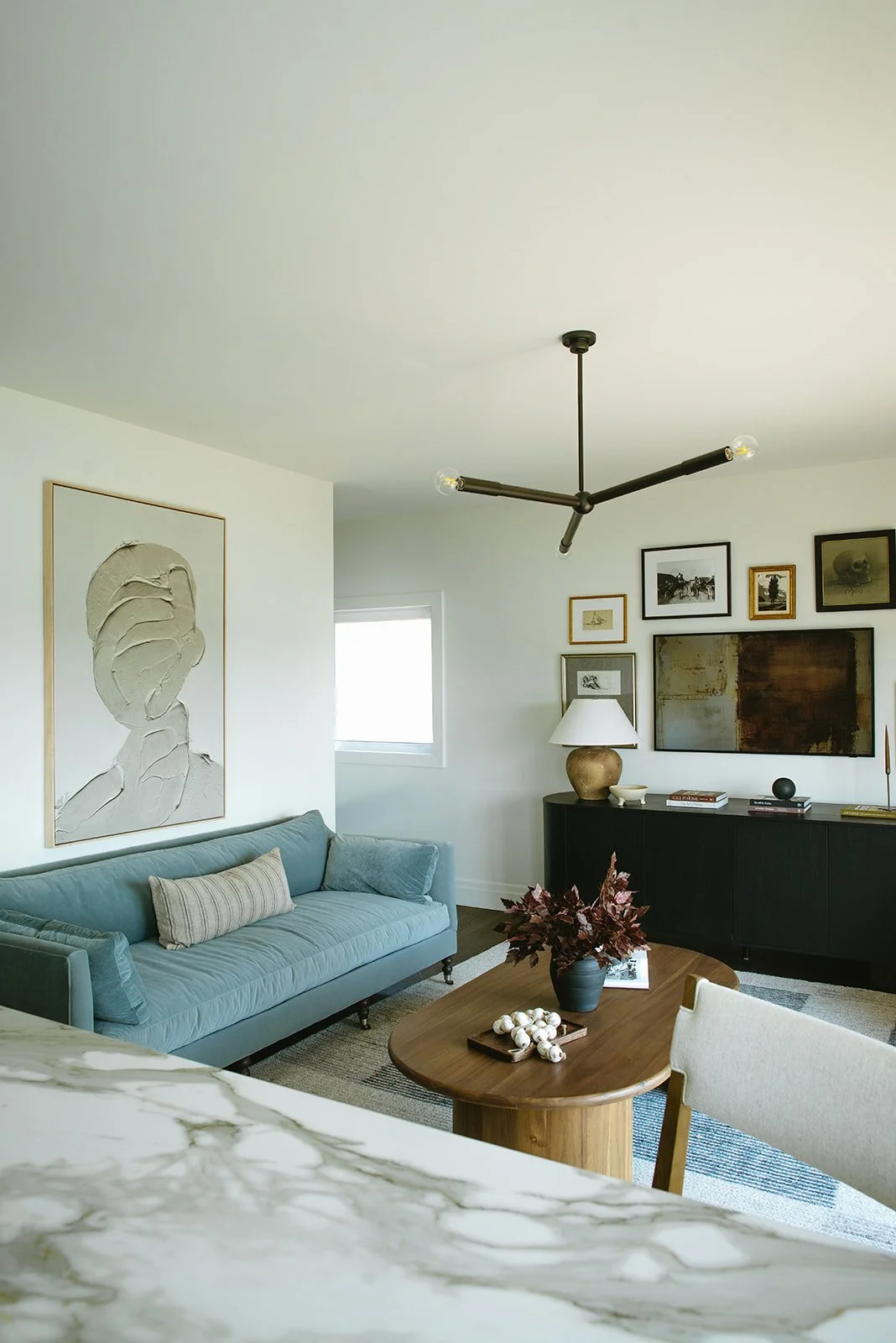 Living room with a blue sofa, abstract wall art, a wooden coffee table with a vase of leaves, assorted books, and decorative objects, and a wall-mounted artwork, lamp, and framed pictures on the wall.