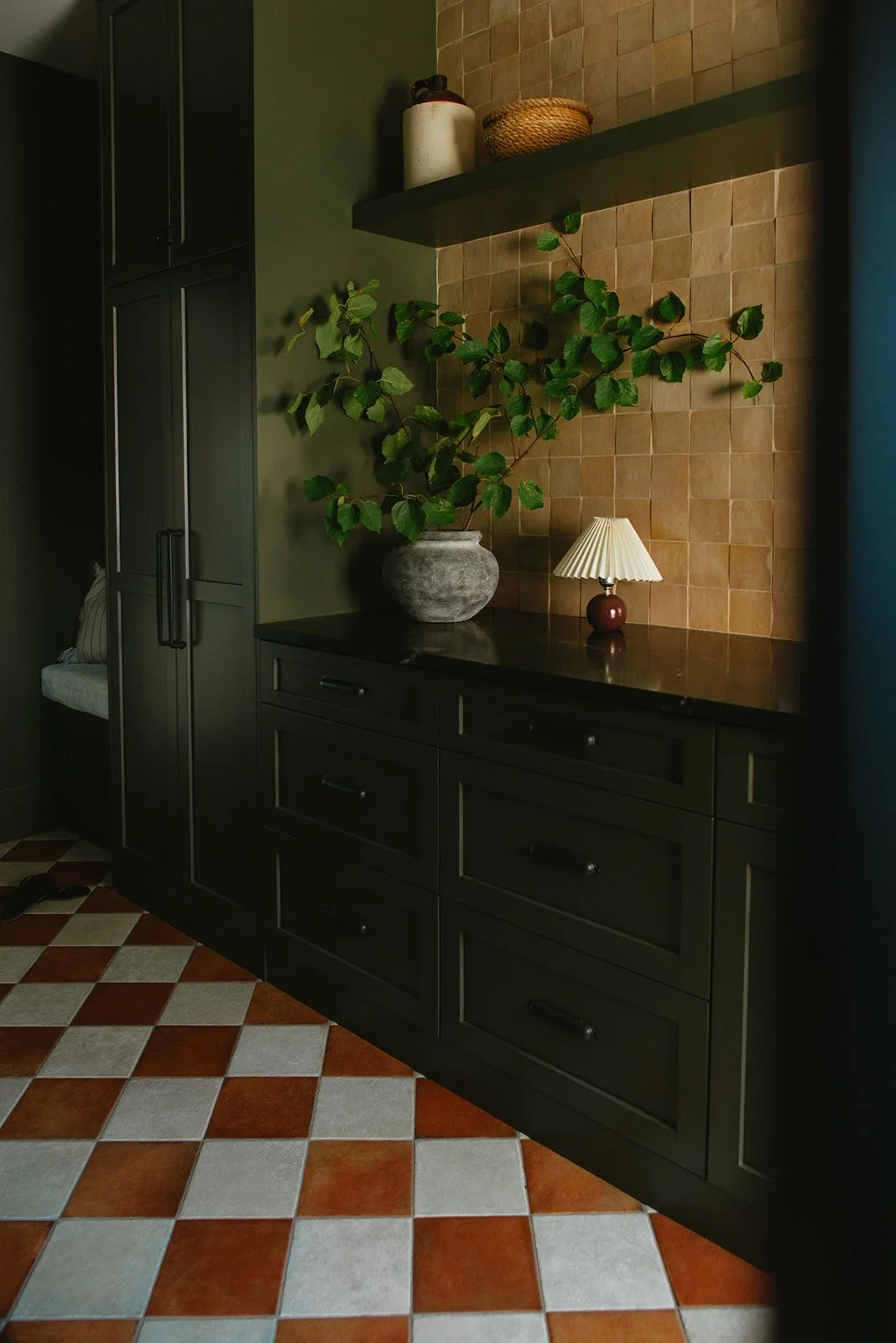 A dark green cabinet and a black countertop with a large potted plant, a small lamp, and shelves with jars on a tiled wall. The floor has red and white checkered tiles.
