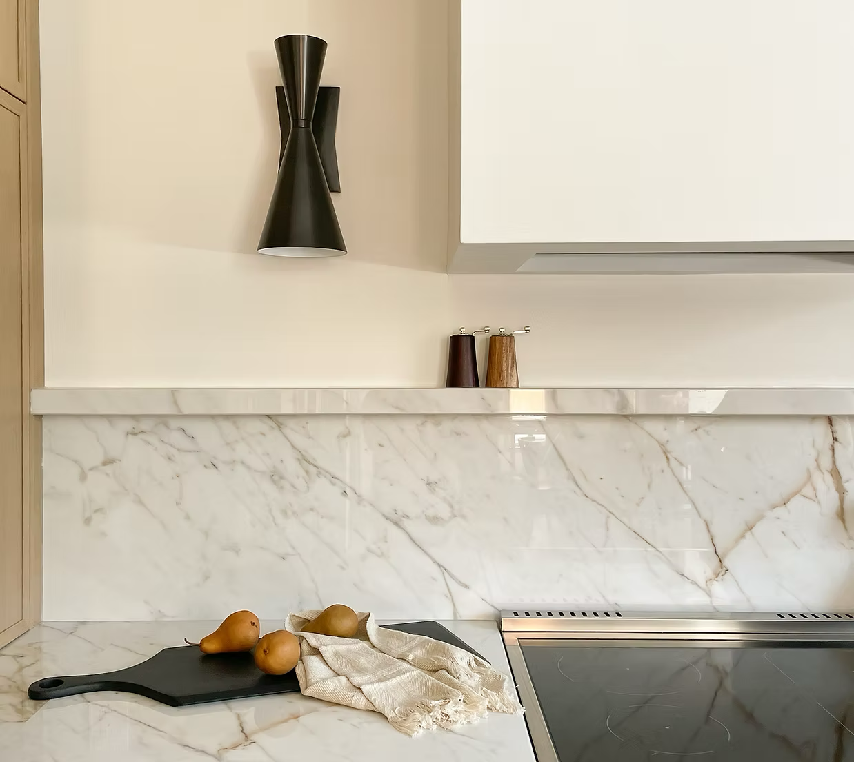 Kitchen counter with three pears on a black cutting board, a cloth, and a stove. Marble backsplash and wall, with wall-mounted black and wood salt and pepper shakers, and a black wall sconce.