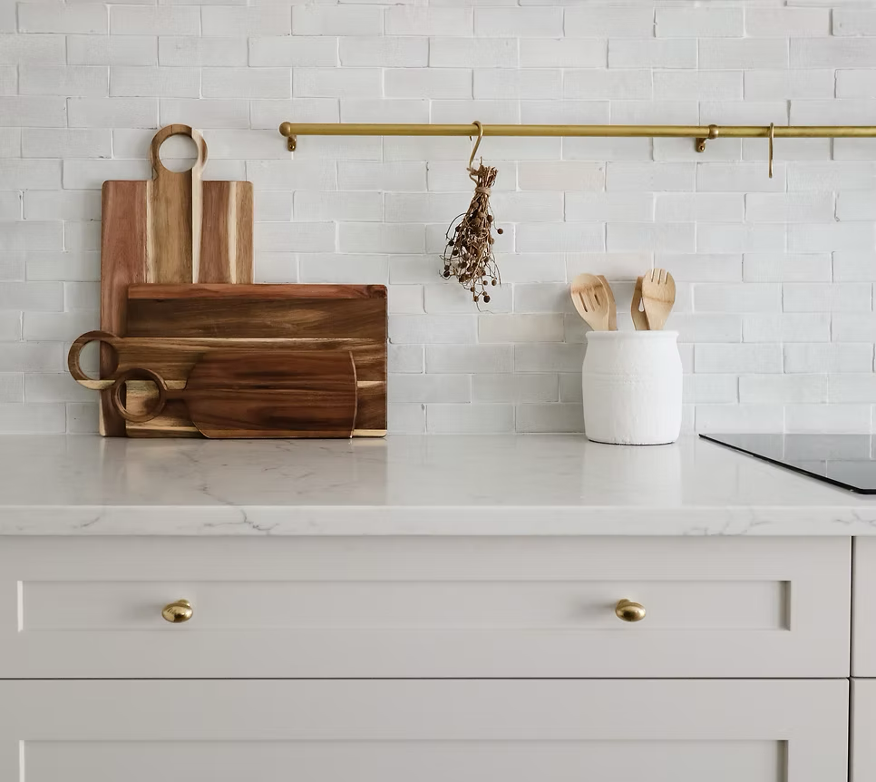 A kitchen countertop with wooden cutting boards, a white container holding wooden utensils, and a dried herb bundle hanging from a brass rod against a white brick wall.