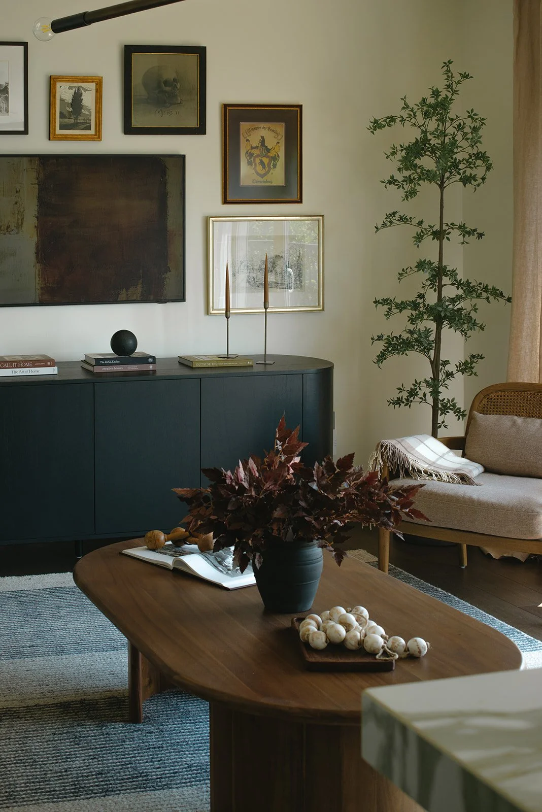 Living room with wall art, a black cabinet, a beige sofa with a throw blanket, a wooden coffee table with a vase of dark leaves, and a tall potted plant.