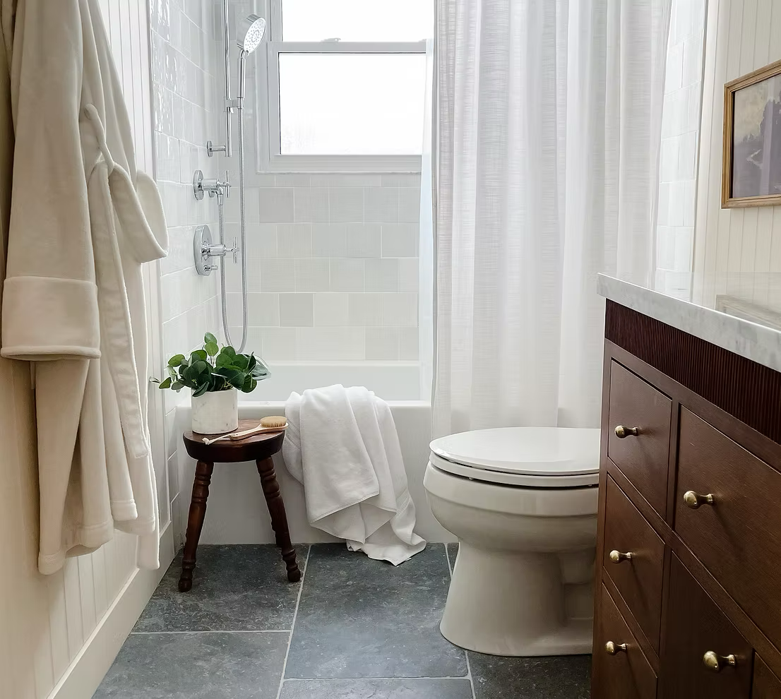Bathroom with a walk-in shower, toilet, wooden cabinet with a marble top, a potted plant on a small wooden stool, towels, and a partially visible framed picture.