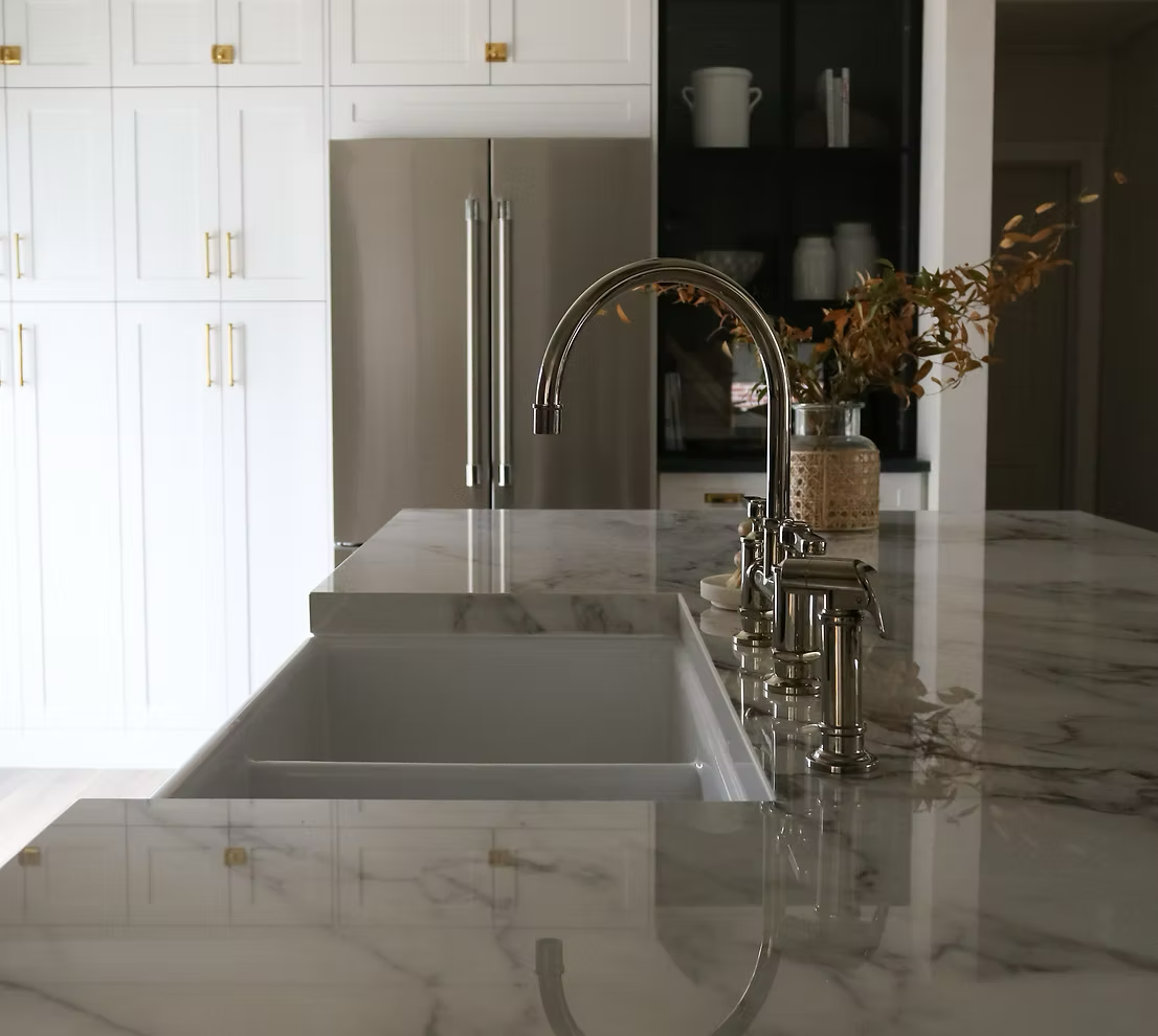 A modern kitchen with a marble countertop, a stainless steel refrigerator, white cabinets with gold handles, and a vase with dried leaves.