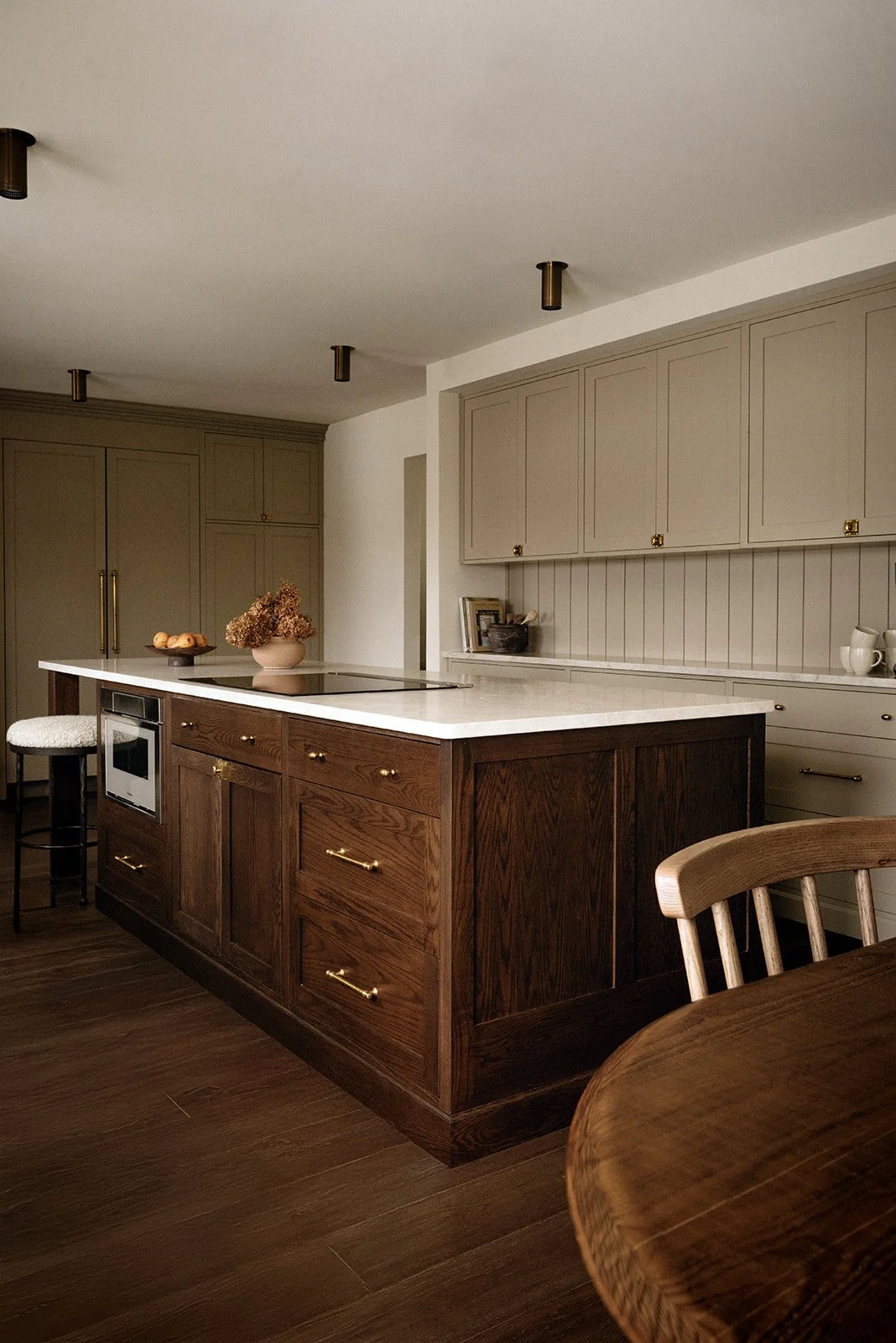 A modern kitchen with beige upper cabinets and dark wood lower cabinets with brass handles, white marble countertops, and a kitchen island with a vase of dried flowers. There is a small oven and a stool with a white cushion at the island. Wooden dini