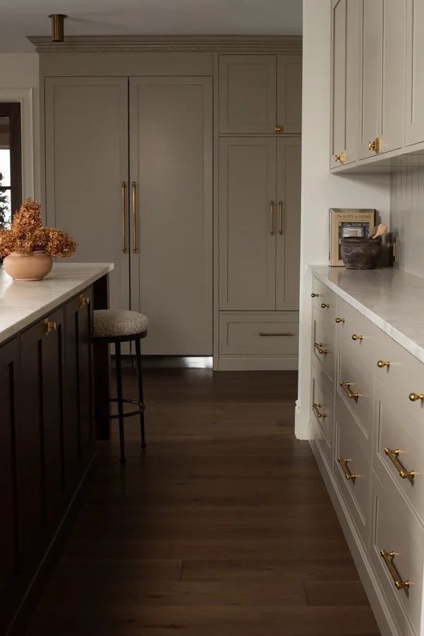 A modern kitchen with white and gray cabinets, gold handles, a white marble countertop, and dark wood flooring. There is a small bar stool next to a dark lower cabinet with dried flowers in a pot on the counter.