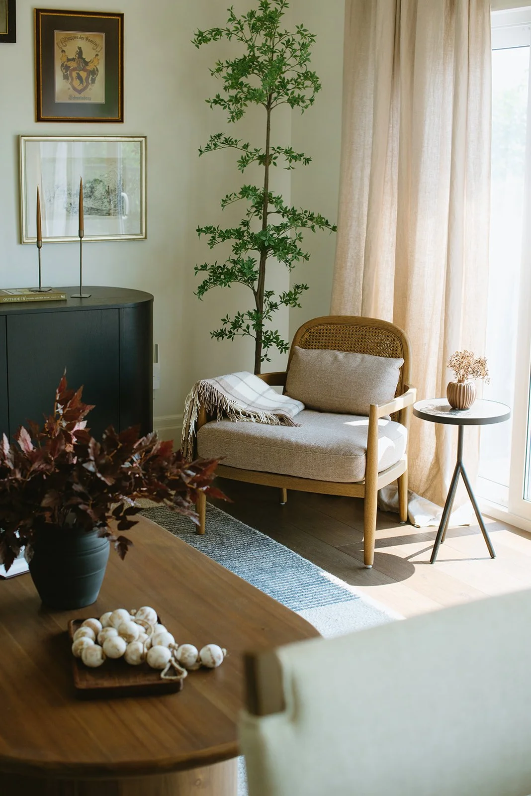 A cozy living room corner featuring a wooden armchair with beige cushions, a small round side table with a flower vase, a tall houseplant, and sunlight streaming through cream-colored curtains.