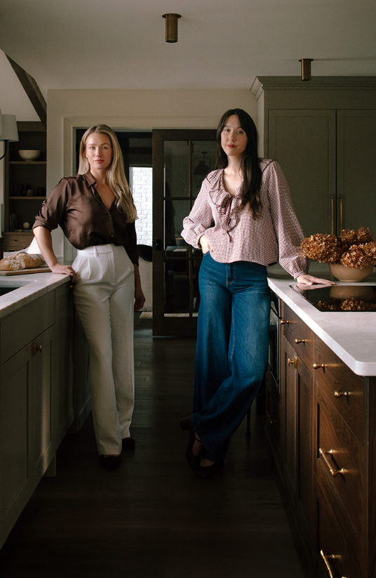 Two women standing in a modern kitchen with dark wood floors, green cabinets, and a white countertop. One woman leans against the counter on the left, wearing a brown blouse and white pants. The other woman stands on the right, wearing a checkered blouse and wide-leg jeans, with her hand resting on the counter.
