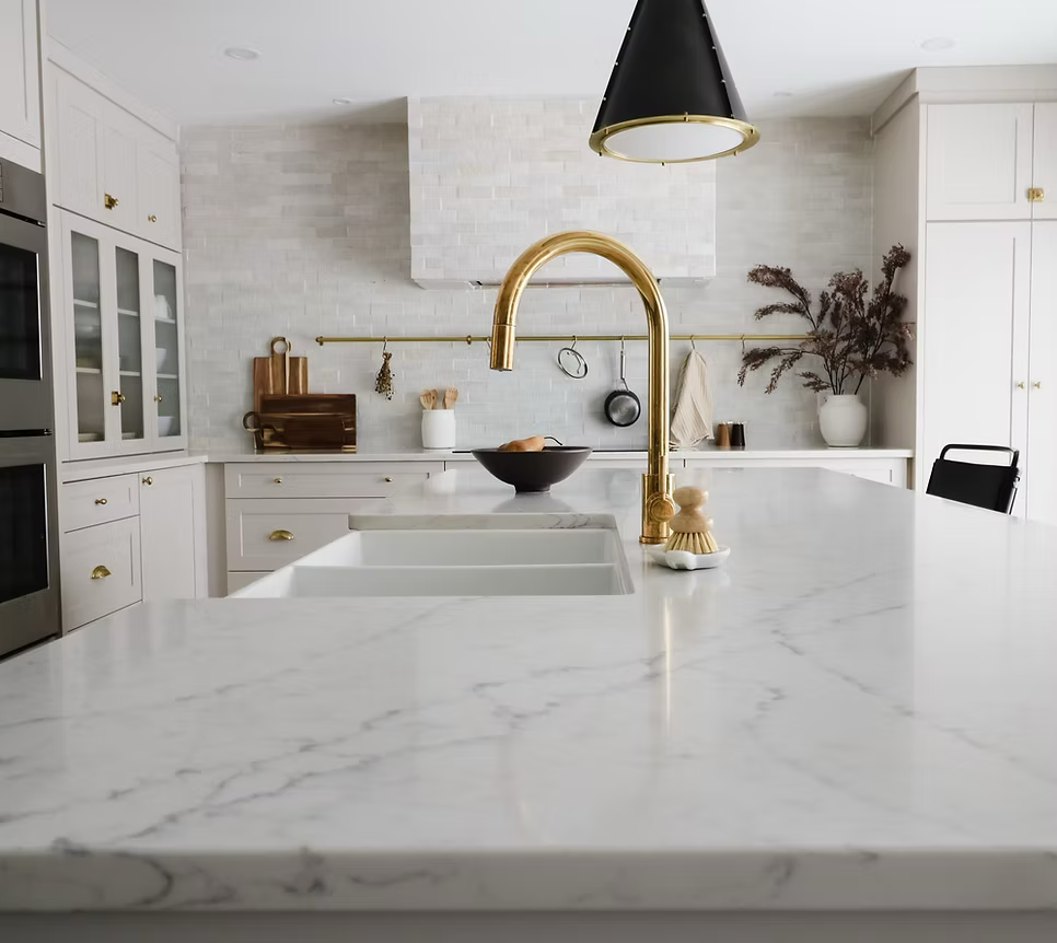 Modern white kitchen with marble countertops, gold faucet, black pendant light, and a black bowl on the counter.
