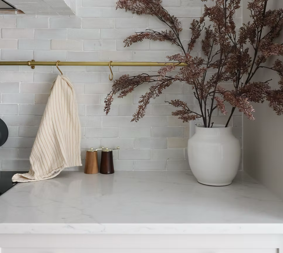 A kitchen countertop with a large white vase containing purple dried branches, two wooden condiment shakers, and a white towel hanging on a brass rod against a white brick wall.