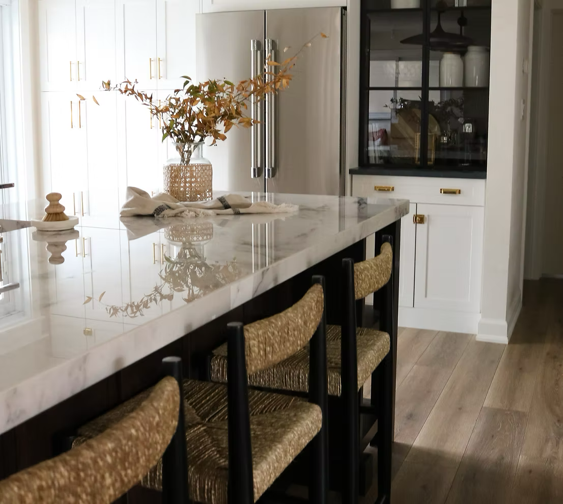 Modern kitchen with white cabinets, marble countertop island, and wood flooring. Decorative vase with leaves on the island, black and gold hardware, and glass-front cabinet with white jars inside.