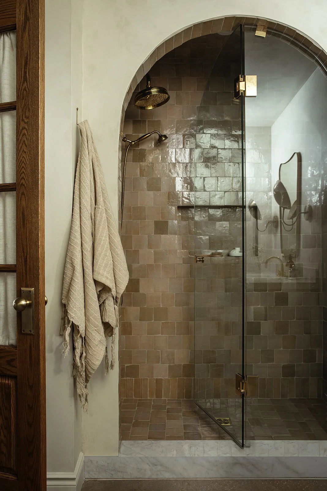 A bathroom with a glass-enclosed shower featuring tan square tiles and a brass rainfall showerhead. Beige towels hang on a hook on a wall next to the shower. The door is wooden with glass panels.